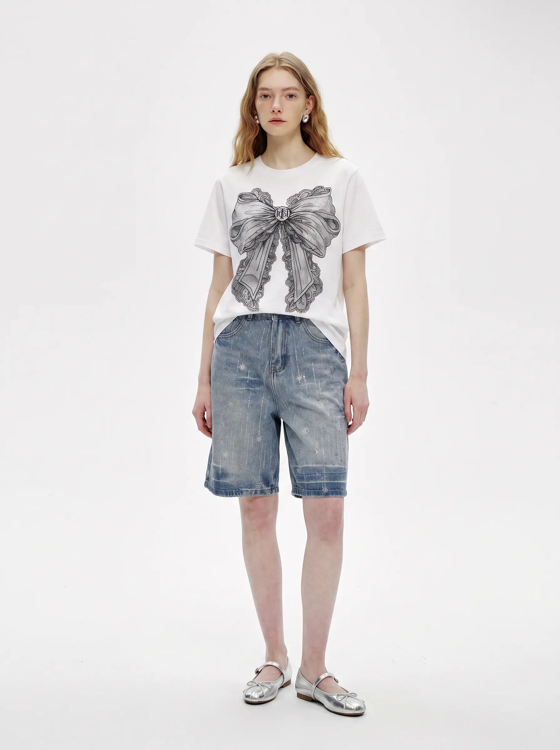 A model stands against a plain light gray studio background wearing a white short-sleeve T-shirt with a large gray bow graphic on the front, light-wash denim shorts, and metallic silver sandals. TCH Rhinestone Vintage Bow Graphic White T-Shirt
