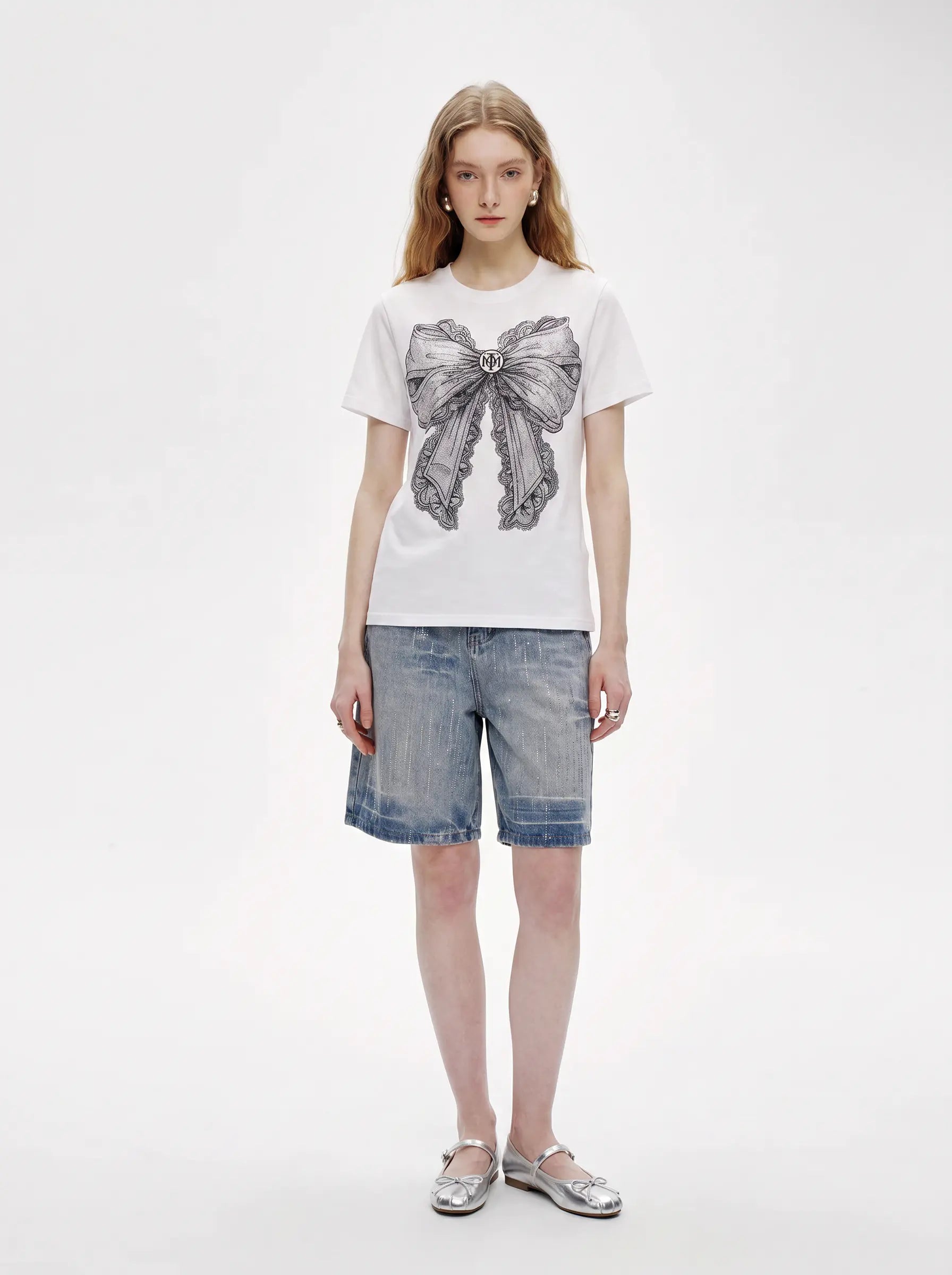 A model stands against a plain white studio background wearing a white short-sleeve T-shirt with a large gray bow graphic on the front, light blue denim shorts, and metallic silver flat sandals. TCH Rhinestone Vintage Bow Graphic White T-Shirt