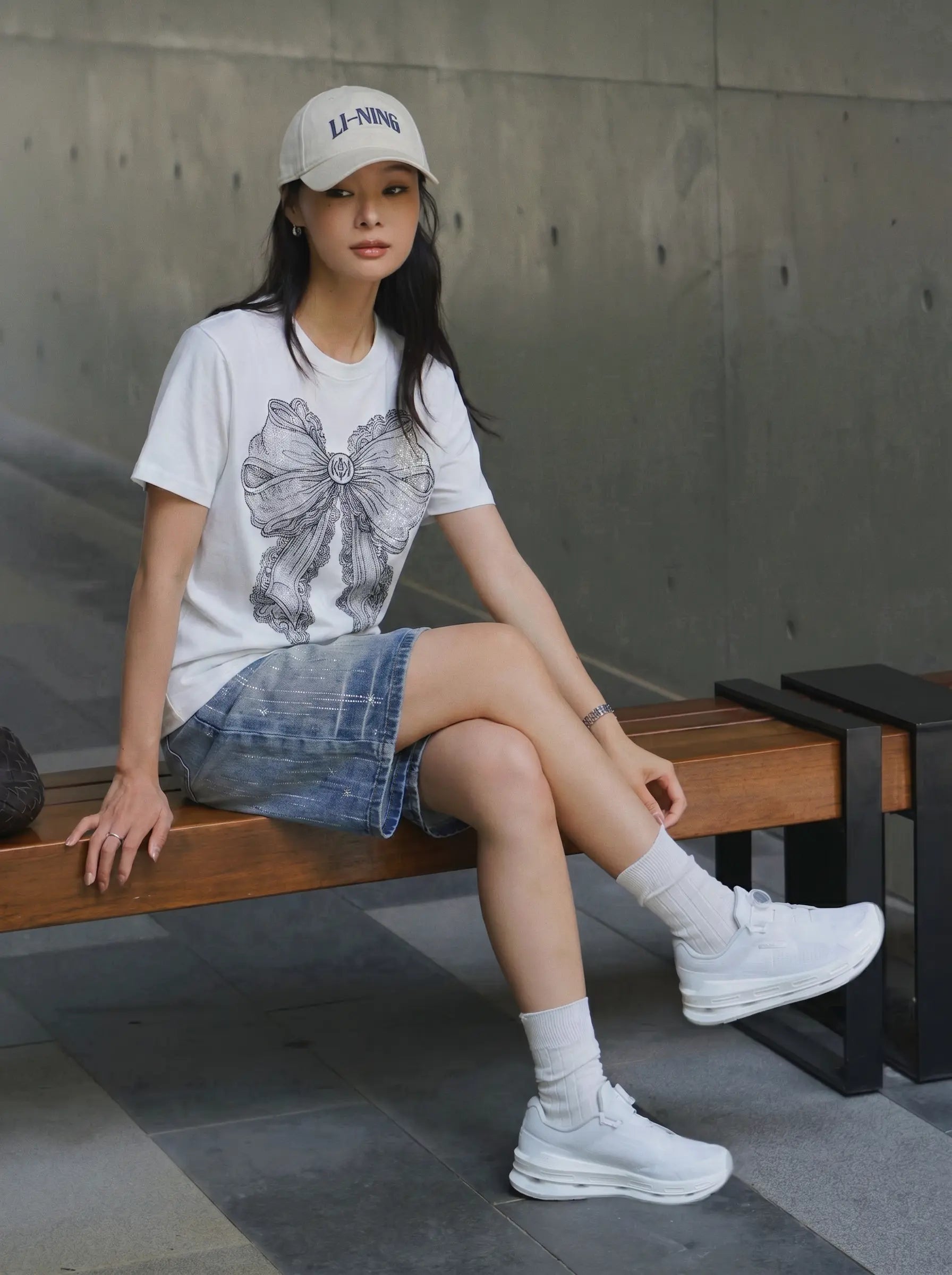 A woman sits on a wooden bench wearing a white graphic T-shirt with a bow design, a denim mini skirt, white socks, and white sneakers. She is also wearing a beige baseball cap with 'LI-NING' printed on it. TCH Rhinestone Vintage Bow Graphic White T-Shirt