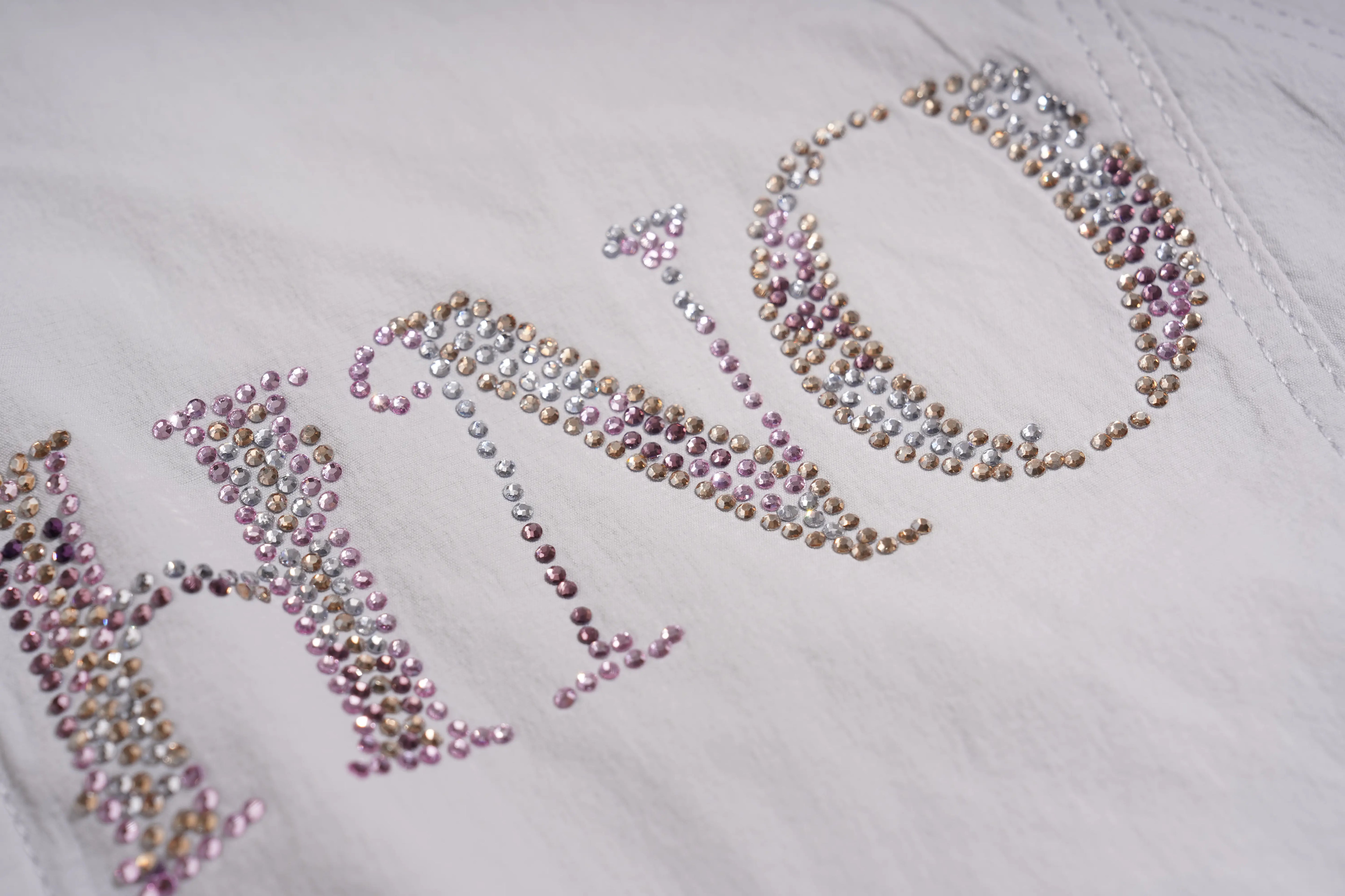 A close-up of a white fabric surface decorated with large rhinestone letters spelling 'HONO' in pink, silver, and gold tones. TCH Rhinestone Wing Active 3-Piece Tracksuit Set