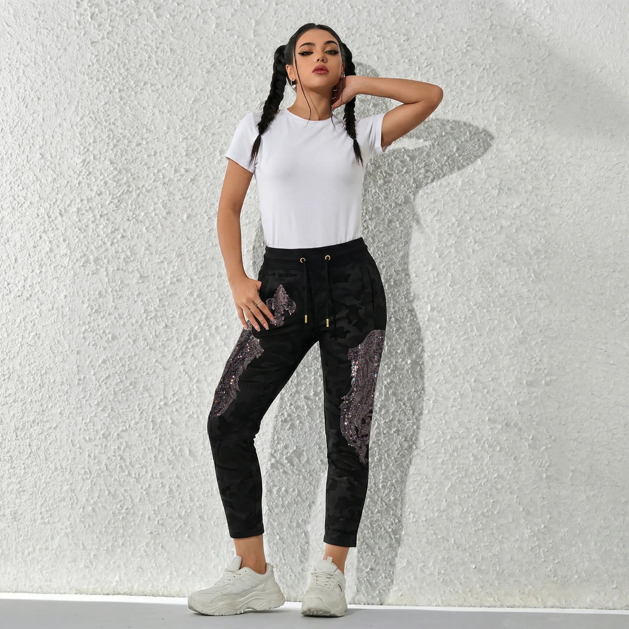Woman wearing a white t-shirt and black sequin pants against a textured white wall. TCH Rhinestone Wing Camo Jogger Pants