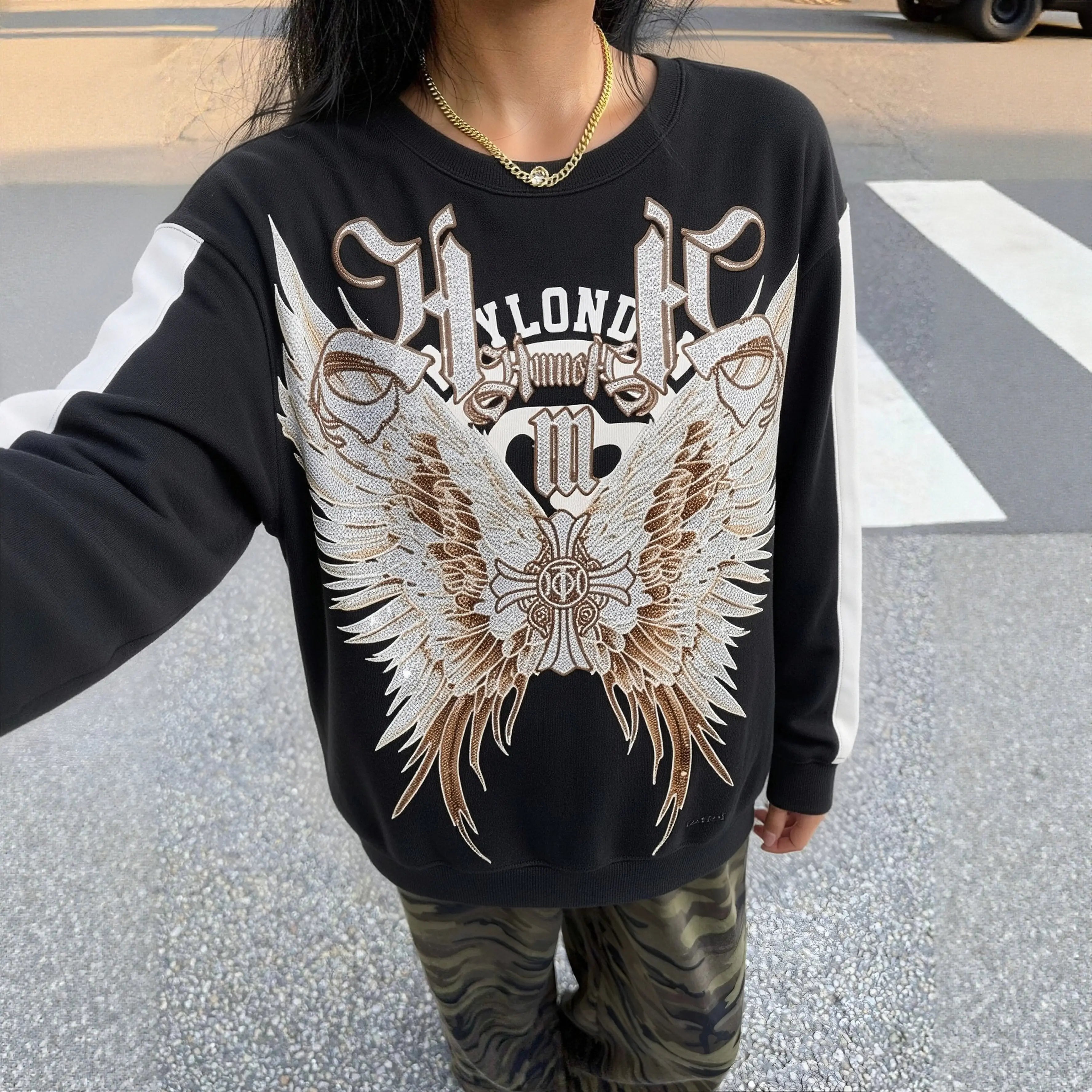 Person wearing a black sweatshirt with a large graphic design on a street. TCH Rhinestone Wing M Embroidered Sweatshirt