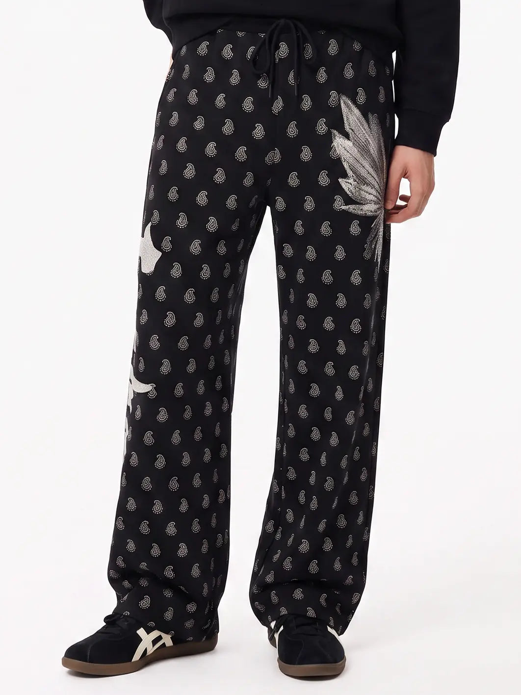 Black wide-leg sweatpants with a small gray paisley print, a large gray leaf applique on the right thigh, and a white graphic on the left leg, modeled with black sneakers against a white studio background. TCH Rhinestone Wing Paisley Wide-Leg Sweatpants