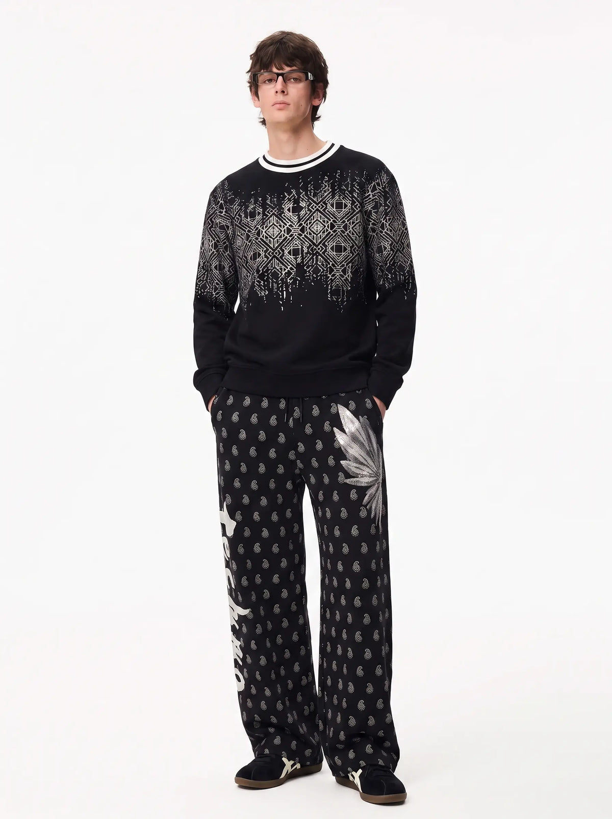A man modeling a black knit crewneck sweater and matching wide-leg trousers with white and gray geometric and floral patterns, set against a plain white studio background. TCH Rhinestone Wing Paisley Wide-Leg Sweatpants