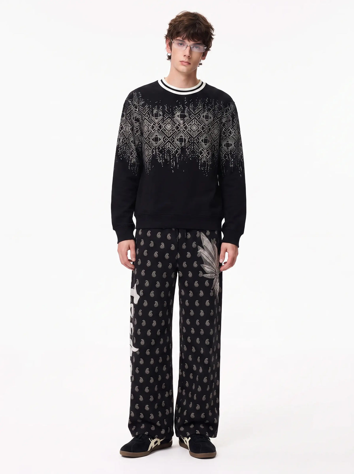 A model wearing a black crewneck sweater with a white geometric pattern across the chest and shoulders, paired with matching black and white patterned trousers, standing against a plain white background. TCH Rhinestone Wing Paisley Wide-Leg Sweatpants