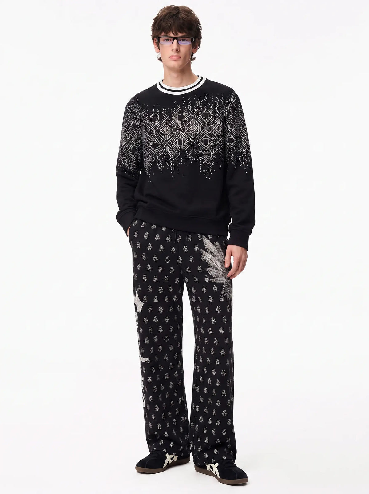 A man modeling a black knit crewneck sweater and matching black patterned trousers with white accents, photographed against a plain white studio background. TCH Rhinestone Wing Paisley Wide-Leg Sweatpants