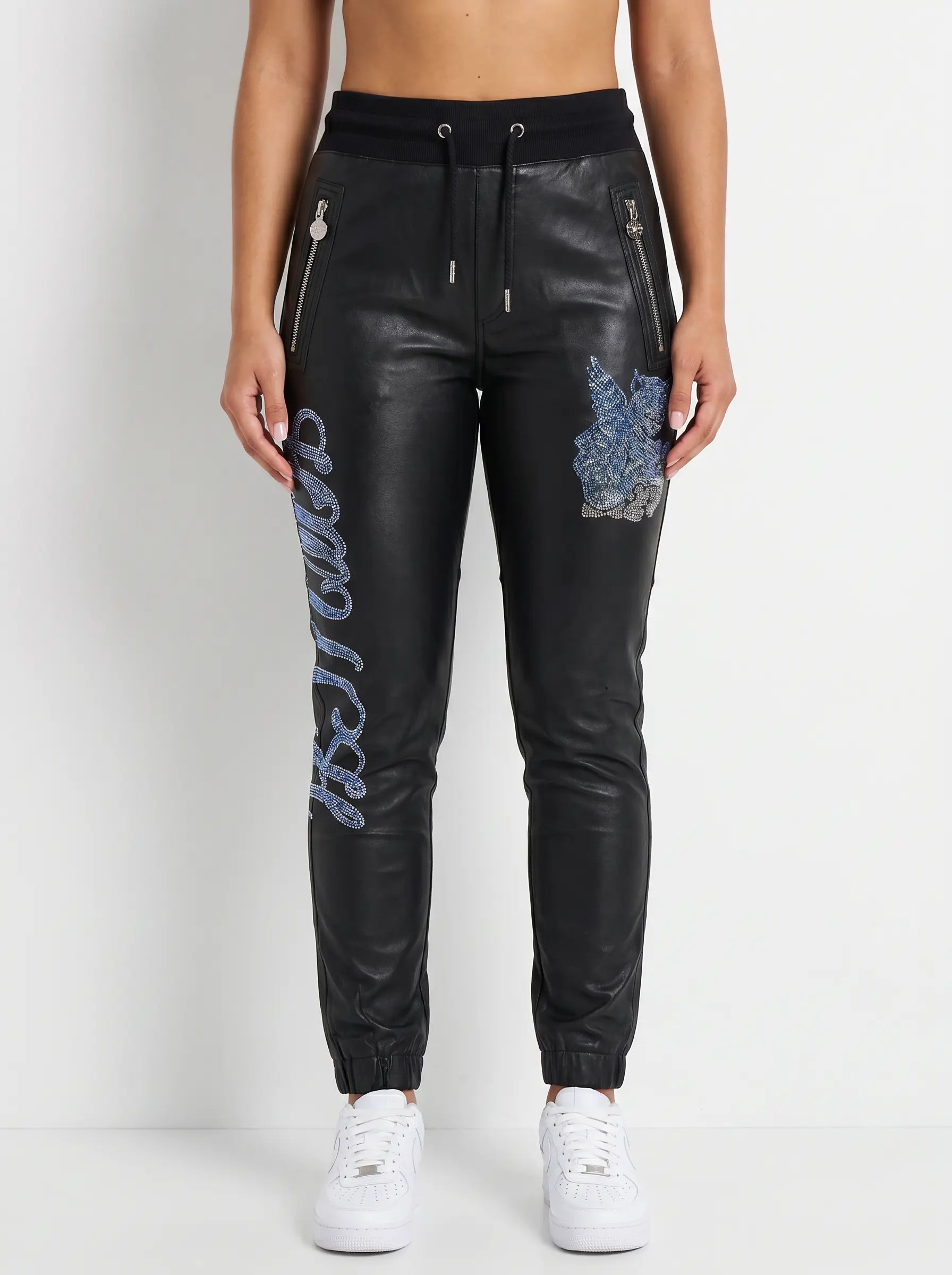 Black faux-leather jogger pants with blue embroidered 'Desire' lettering down one leg, a blue floral patch on the thigh, and silver zipper details, modeled with white sneakers against a white studio background. TCH Rhinestone Wing Teddy Faux Leather Jogger Pants