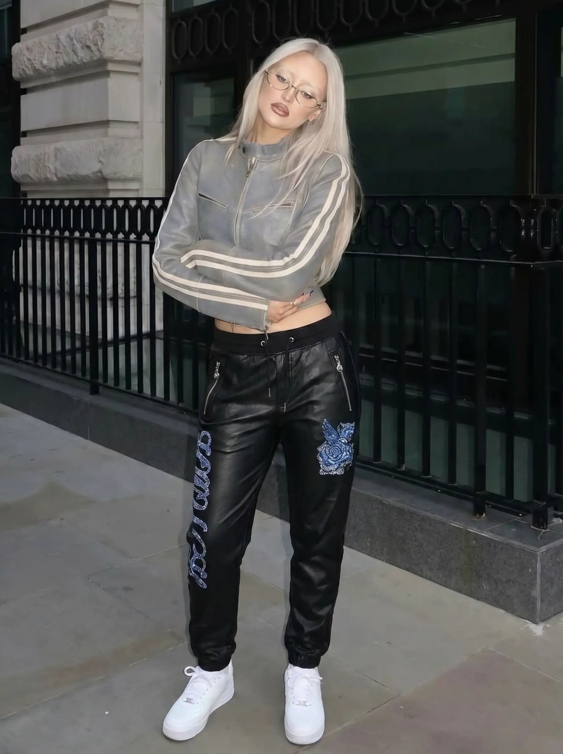 A woman stands outdoors in front of a building with a black metal railing, wearing a sheer gray zip-up jacket with white stripes, black leather jogger pants with blue graphics, and white sneakers. TCH Rhinestone Wing Teddy Faux Leather Jogger Pants