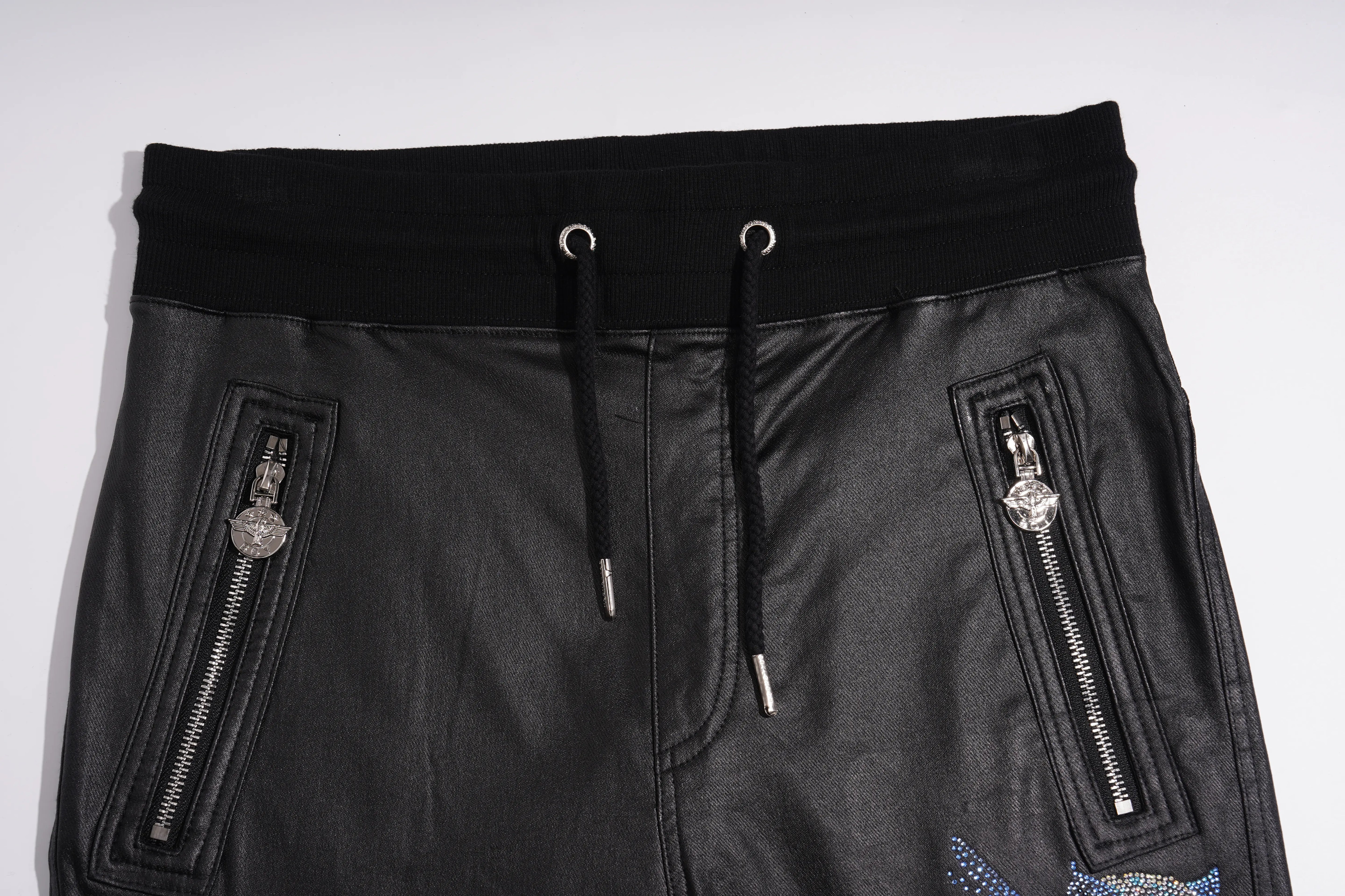 Black faux leather pants featuring a wide black elastic waistband with black drawstrings, two vertical zippered pockets with silver-tone hardware, and a small embroidered patch near the bottom right. TCH Rhinestone Wing Teddy Faux Leather Jogger Pants