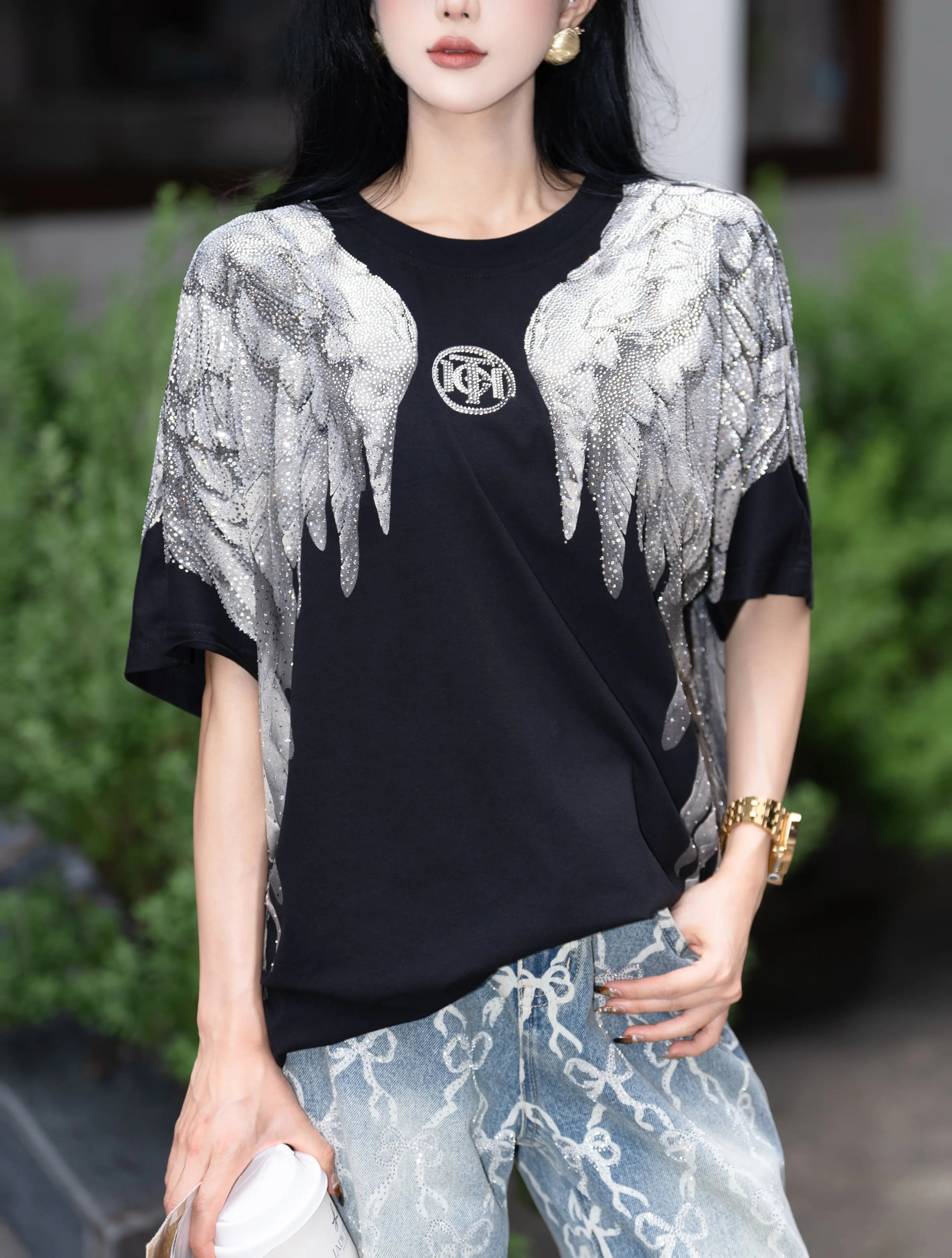 Person wearing a black t-shirt with silver angel wings and a logo, standing outdoors. TCH Rhinestone Wings Logo Black Graphic T-Shirt