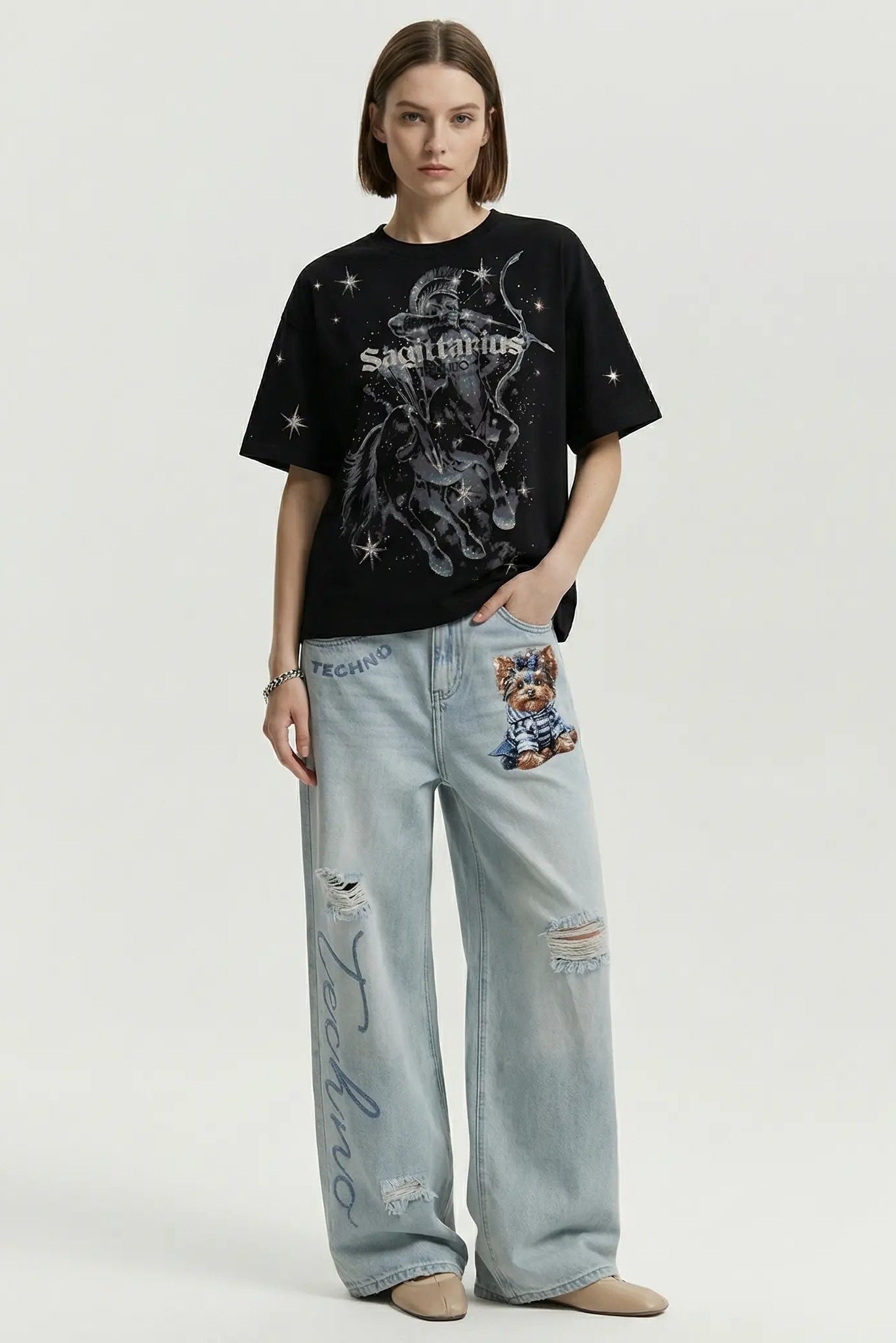 A model stands against a light gray studio background wearing a black graphic T-shirt with a white celestial print and light-wash distressed jeans with decorative patches and embroidery. TCH Sagittarius Celestial Graphic Oversized T-Shirt