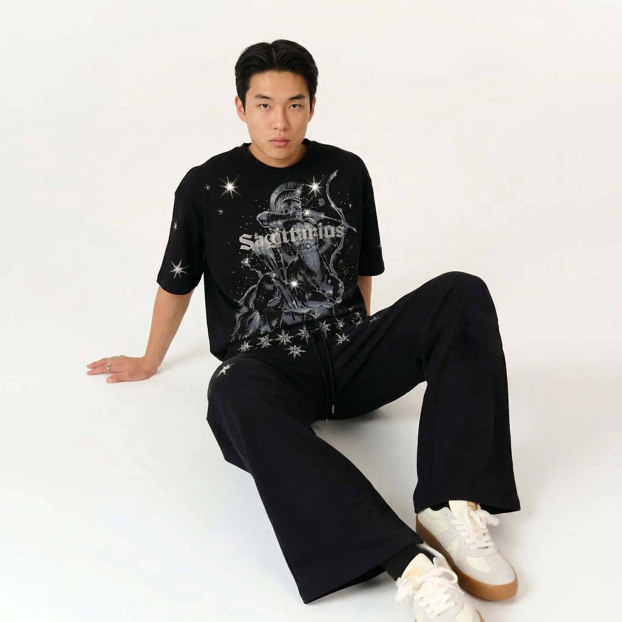 A man wearing a black graphic t-shirt with a silver and white skull design and the word 'Skalpers' on the front, paired with black wide-leg pants and white sneakers, sitting against a plain white studio background. TCH Sagittarius Celestial Graphic Oversized T-Shirt