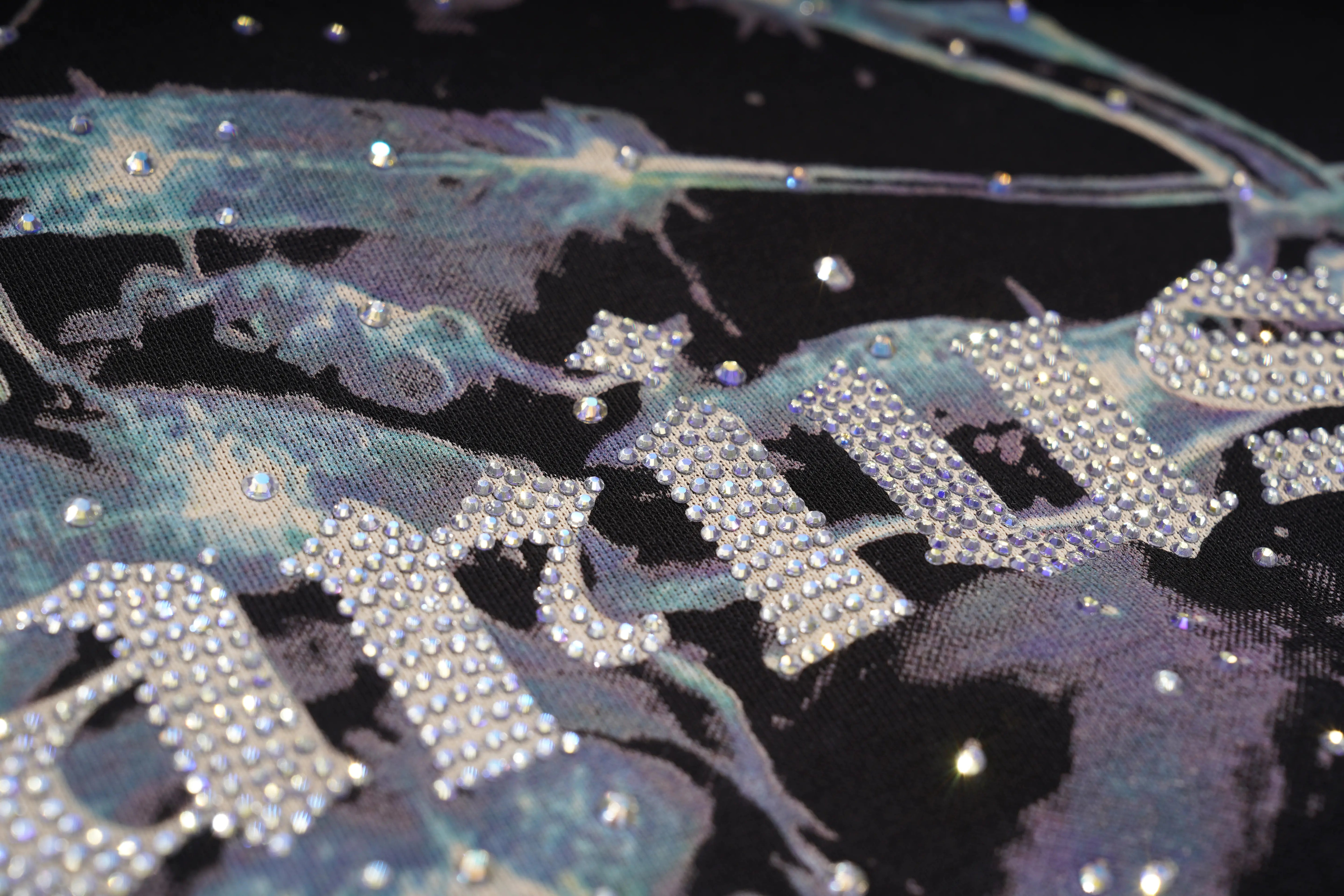 Close-up of a black fabric garment embellished with iridescent blue-green abstract print and large silver rhinestone lettering. TCH Sagittarius Celestial Graphic Oversized T-Shirt