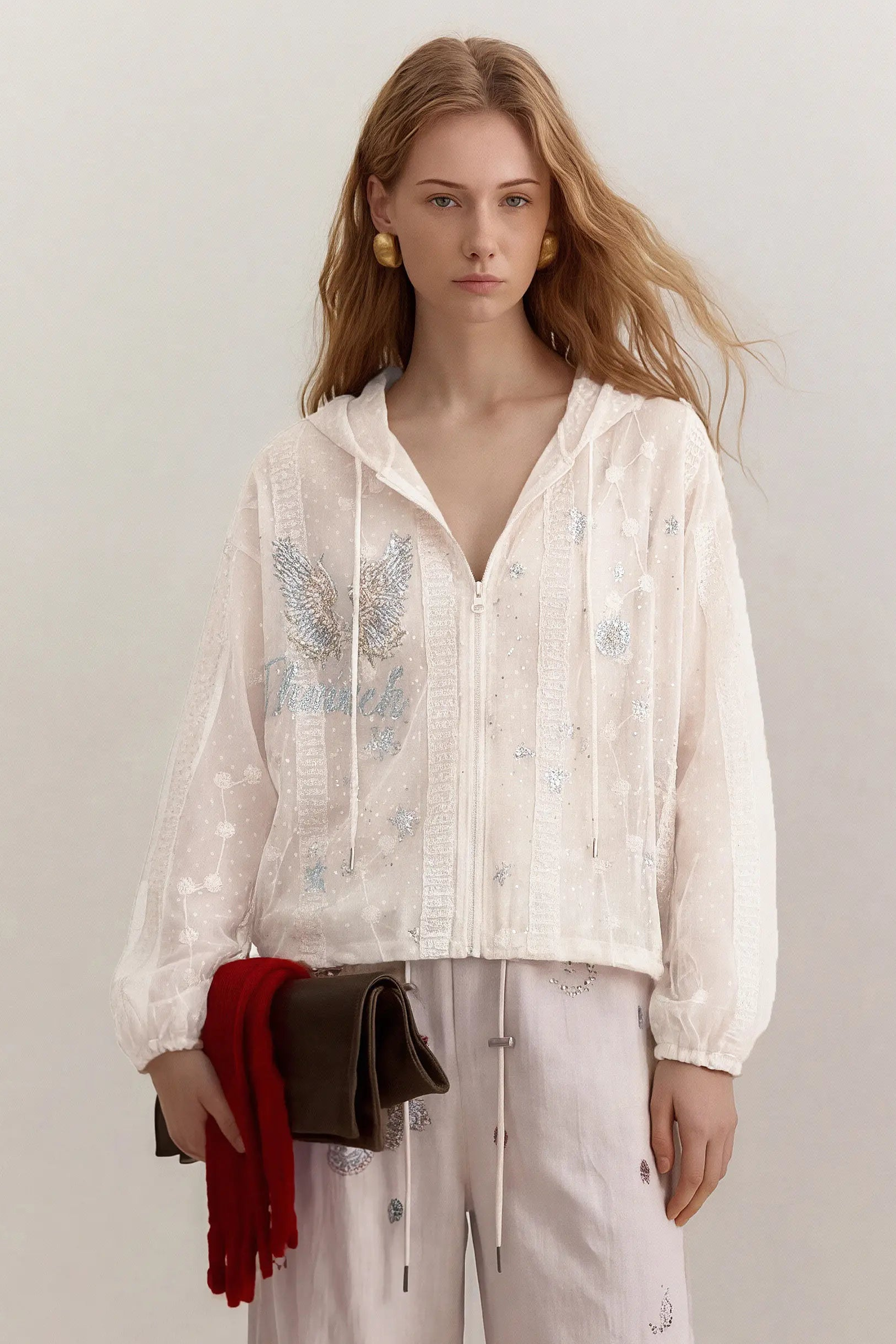 A model wearing a white hooded zip-up jacket with delicate blue floral embroidery and sequins, paired with white pants and holding a red fringed scarf and a dark brown clutch bag, set against a plain light gray studio background. TCH Sheer Rhinestone Angel Star Zip-Up Hoodie