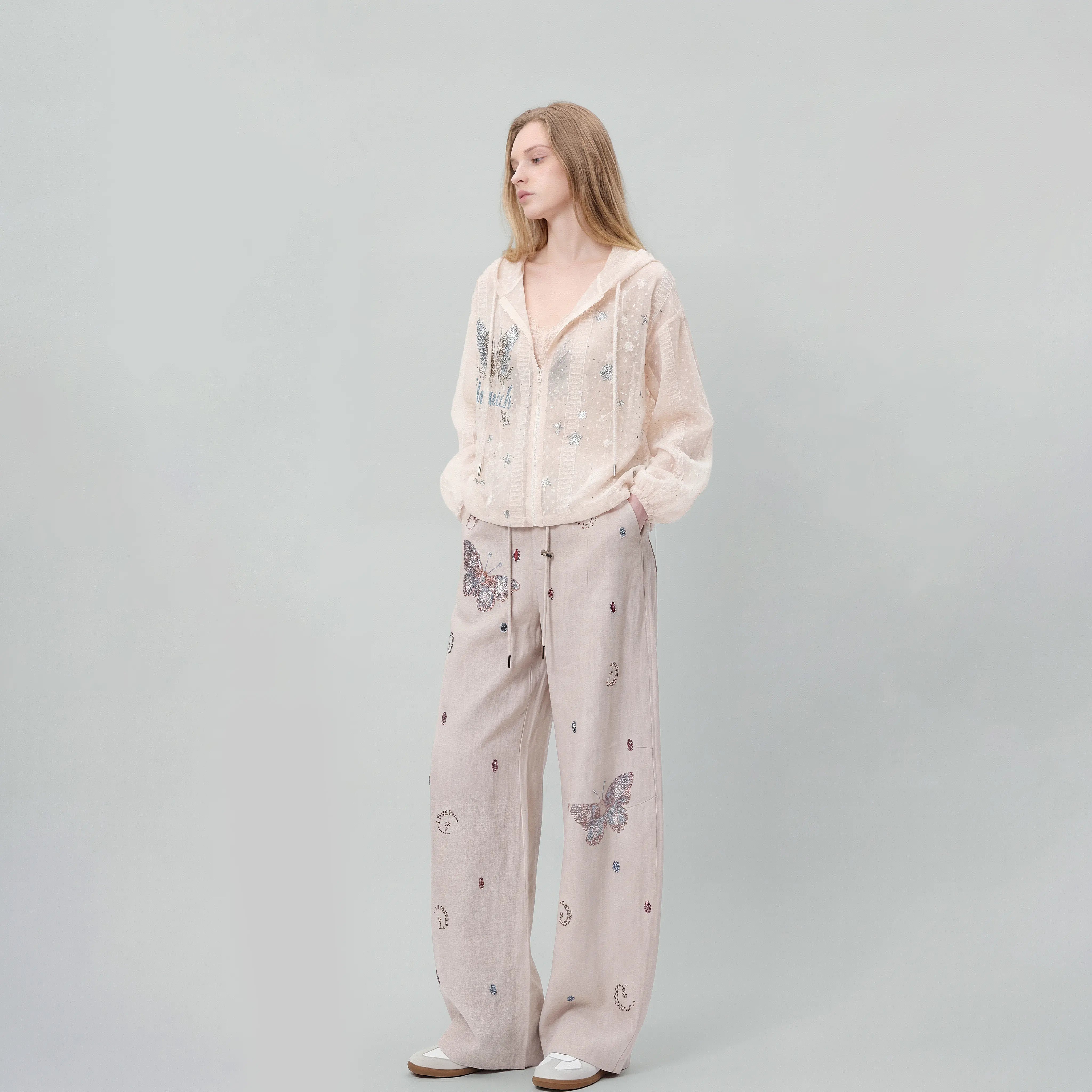 A woman stands against a light gray studio background wearing a matching lounge set consisting of a sheer long-sleeve hooded top and wide-leg pants, both in a pale blush pink color with butterfly and moon prints. TCH Sheer Rhinestone Angel Star Zip-Up Hoodie