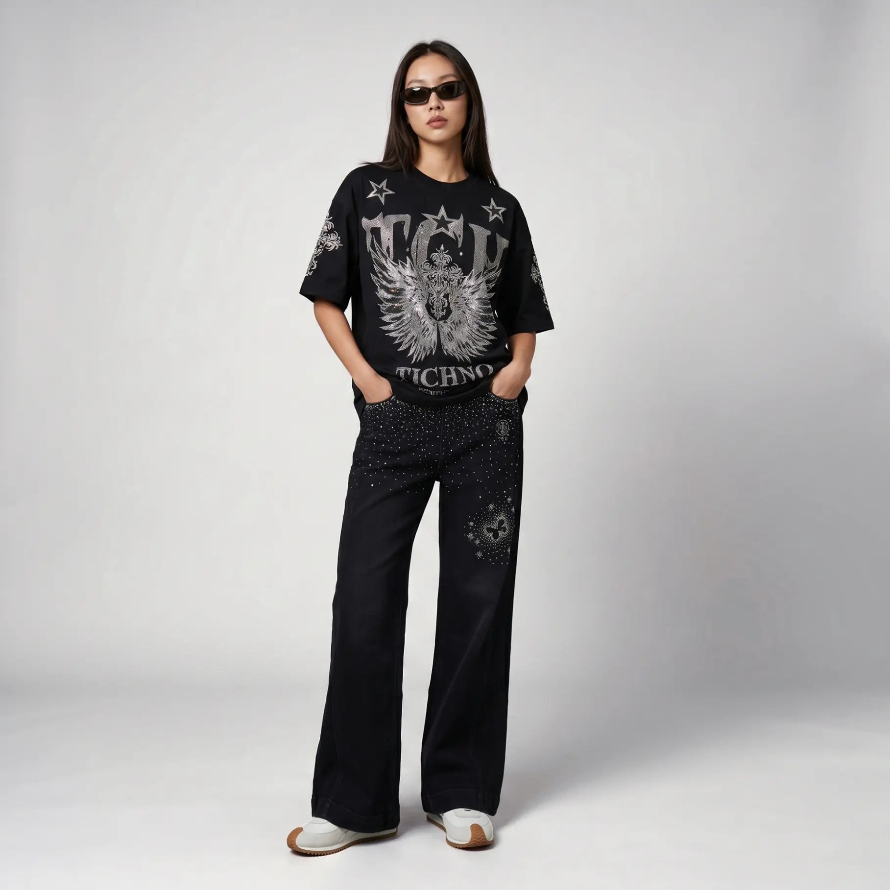 A model stands against a light gray studio background wearing a black graphic T-shirt with a large white angel design and the word 'Techno', paired with black wide-leg pants and white sneakers. TCH Silver Wing Techno Oversized Graphic T-Shirt