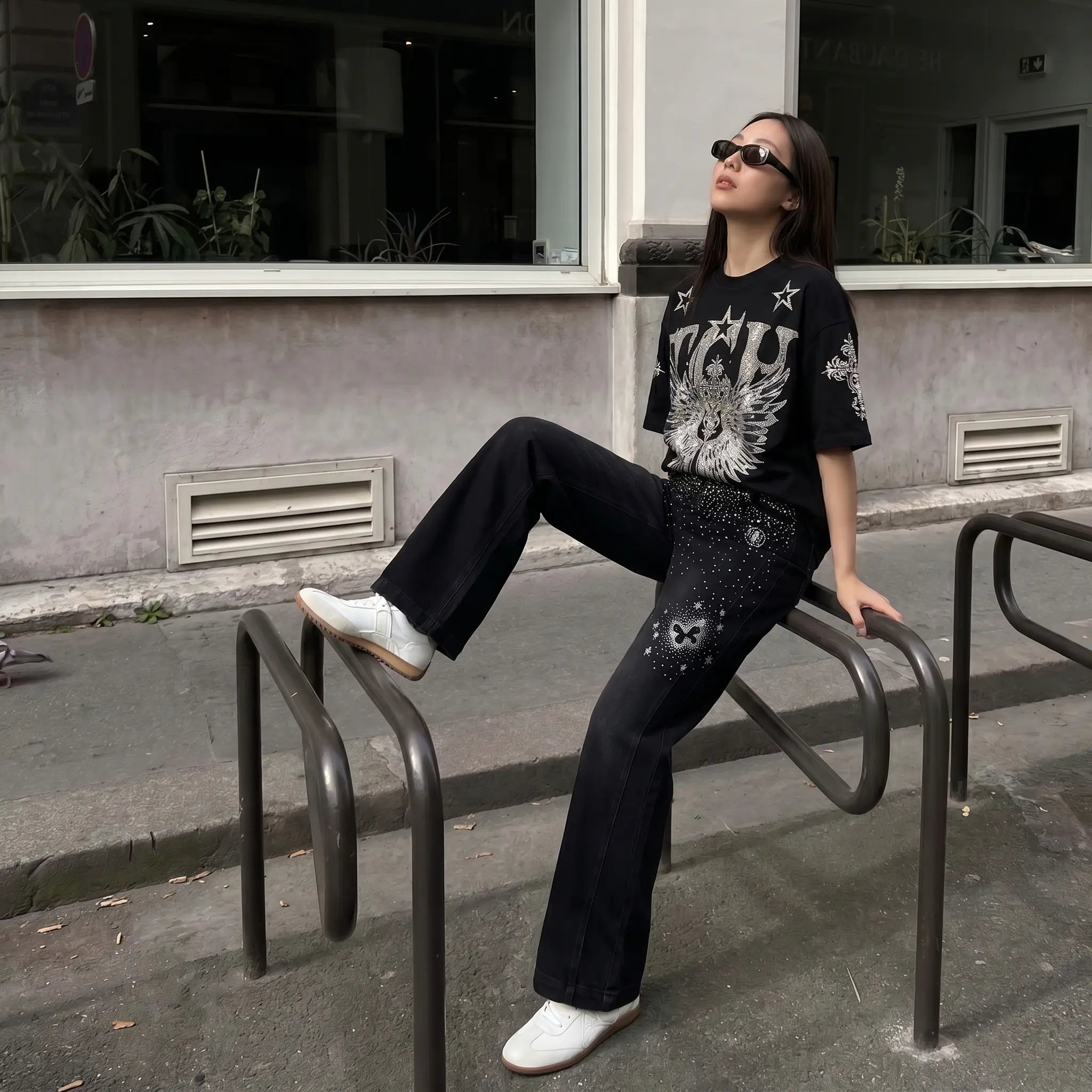 A woman wearing a black graphic t-shirt and black embellished flare pants sits on a metal bike rack outdoors in front of a building. TCH Silver Wing Techno Oversized Graphic T-Shirt