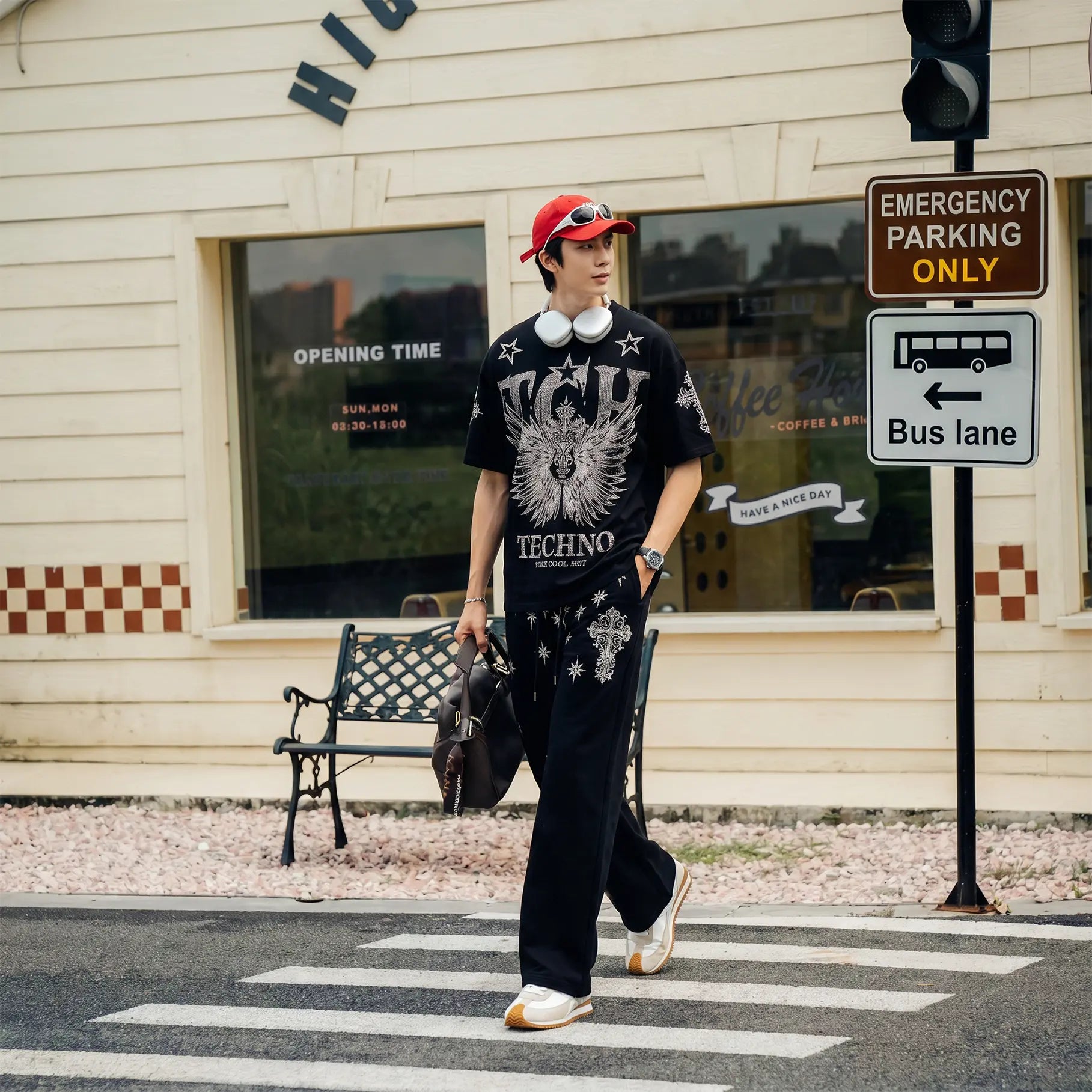 A man walking across a street crosswalk wearing a black graphic t-shirt with 'VHS TECHNO' and a wing design, black pants, a red cap, and white headphones around his neck. The background shows a beige building with a window, a bench, and a 'Bus lane' sign. TCH Silver Wing Techno Oversized Graphic T-Shirt