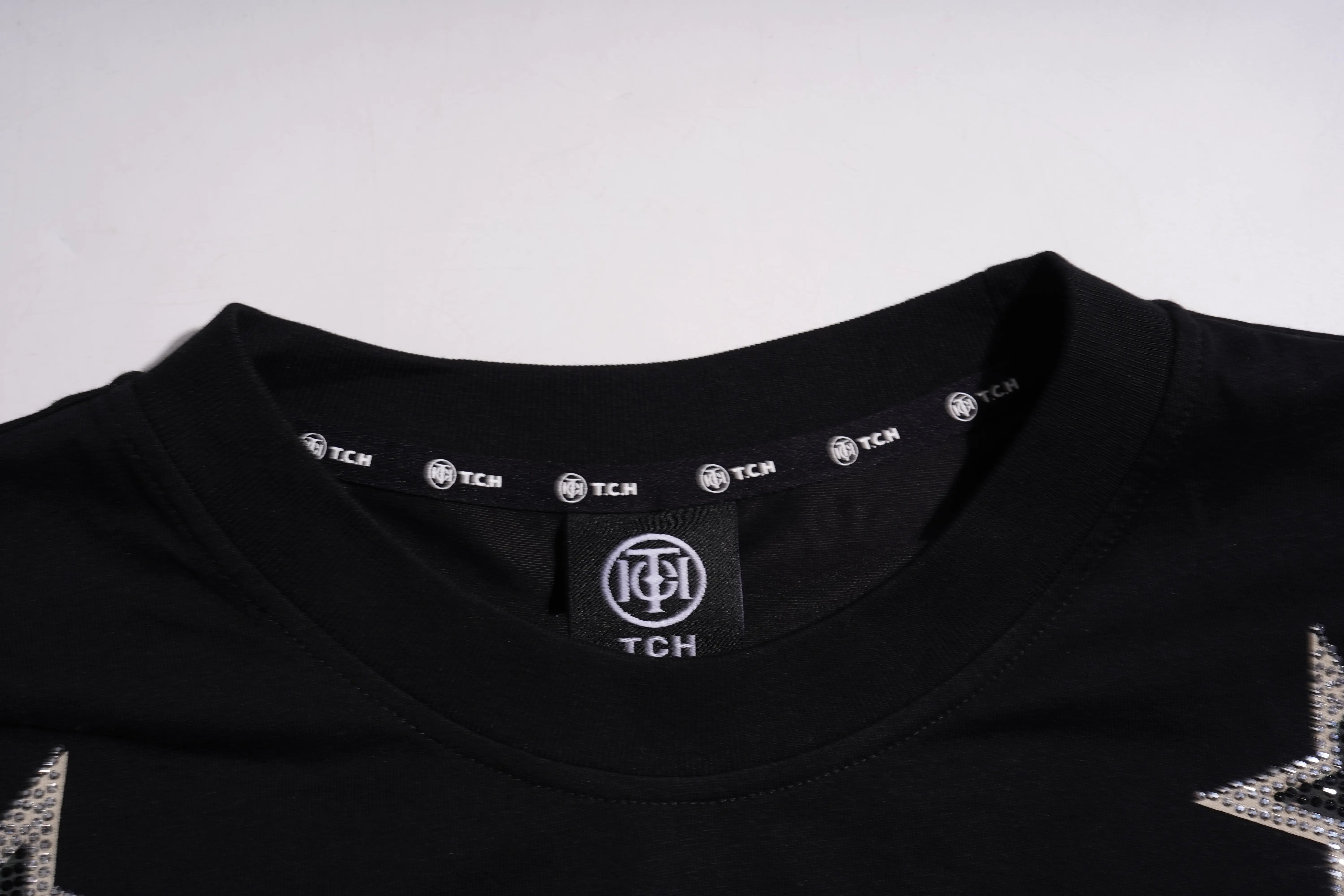Close-up of a black crewneck t-shirt showing the inner neck label with 'TCH' branding, silver graphic details on the shoulders, and a light gray background. TCH Silver Wing Techno Oversized Graphic T-Shirt