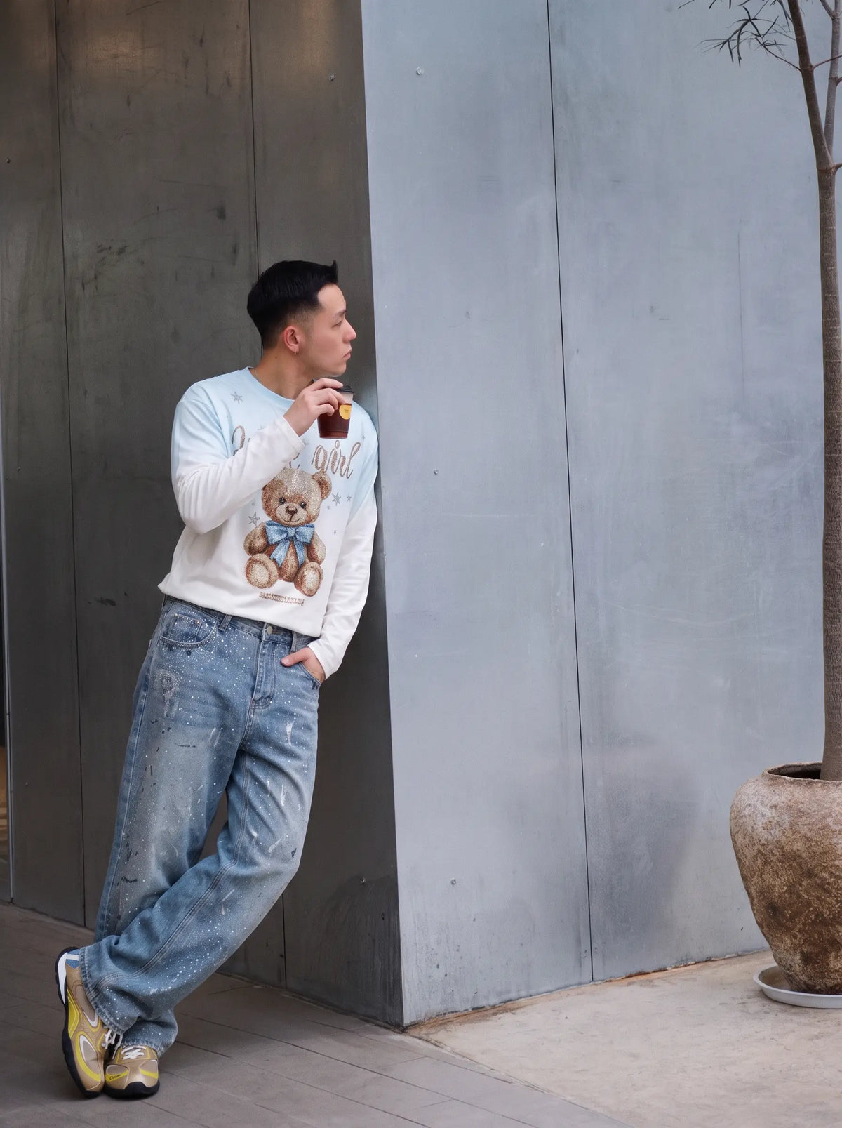 A man wearing a white long-sleeve crewneck sweatshirt with a teddy bear graphic and light wash blue jeans, leaning against a gray concrete wall while holding a drink. TCH Small Bear Angel Wings Rhinestone Graphic White Long Sleeve T-Shirt