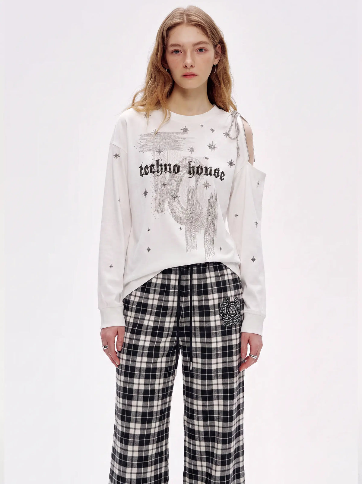 A model wearing a white long-sleeve sweatshirt with a gothic-style graphic print that reads 'techno house' and black-and-white plaid wide-leg pants, photographed against a plain white studio background. TCH Starry Western Graphic White Long Sleeve Top