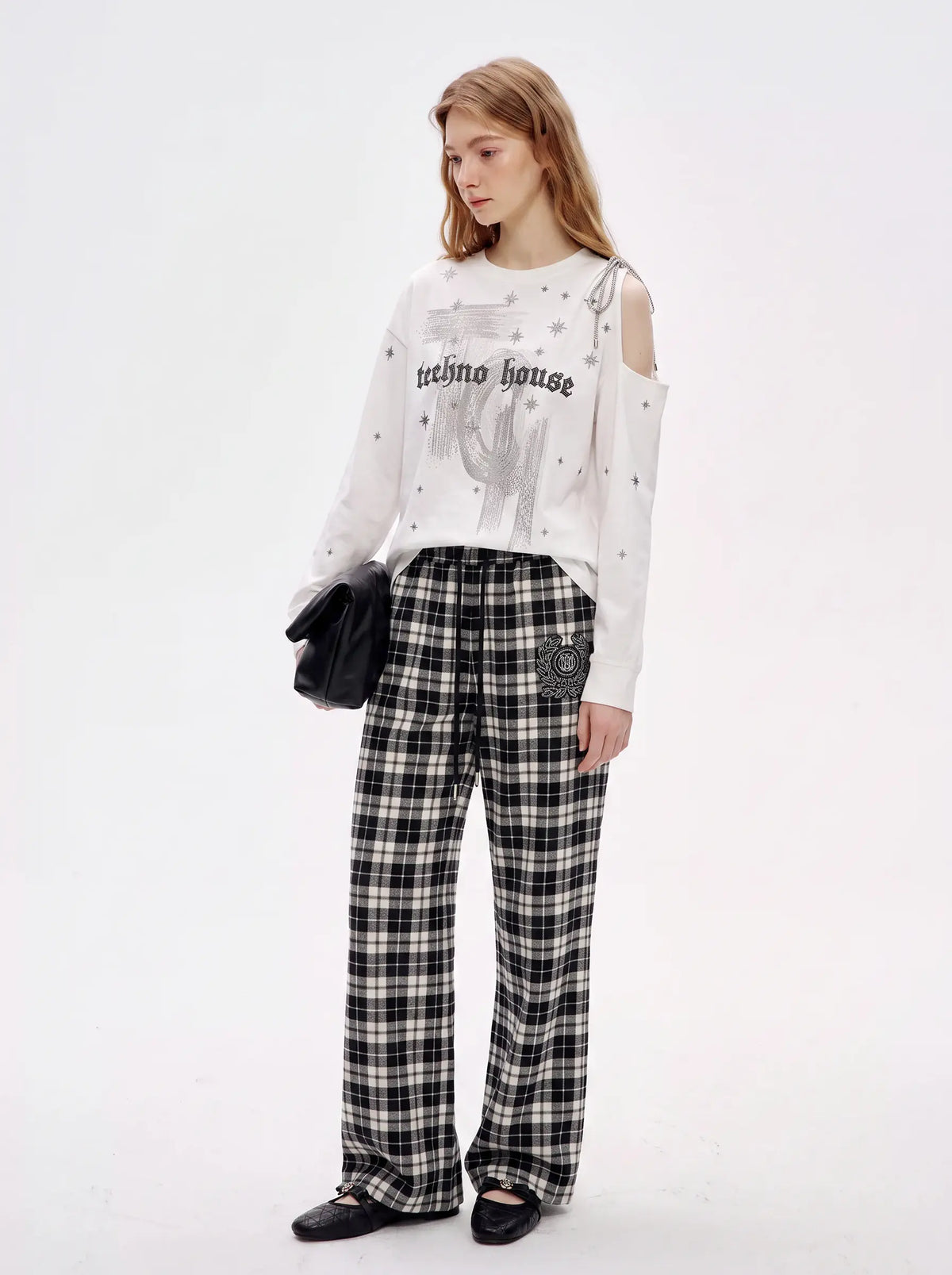 A model stands against a plain white studio background wearing a white long-sleeve top with a graphic print and rhinestone embellishments, paired with black-and-white plaid wide-leg pants and black flats. She holds a small black handbag. TCH Starry Western Graphic White Long Sleeve Top