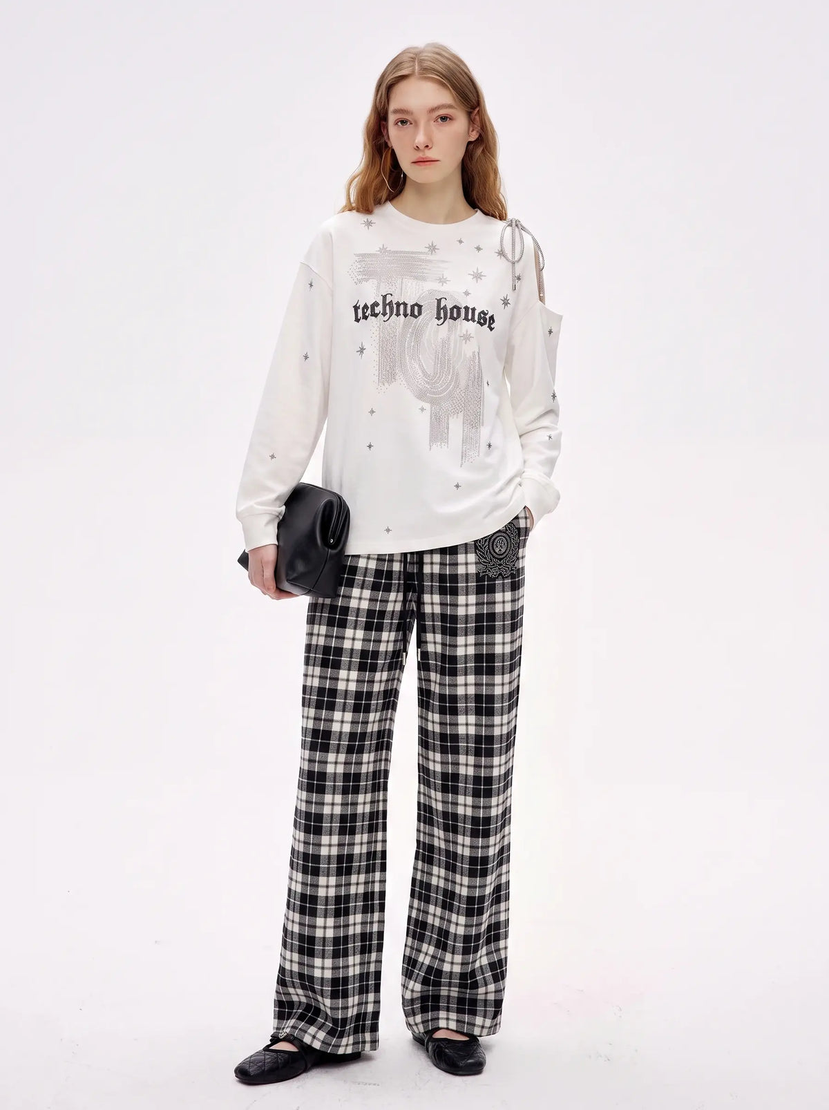 A model stands against a plain white studio background wearing a white graphic sweatshirt with 'techno house' text, black-and-white plaid wide-leg pants, and black shoes, while holding a small black clutch. TCH Starry Western Graphic White Long Sleeve Top