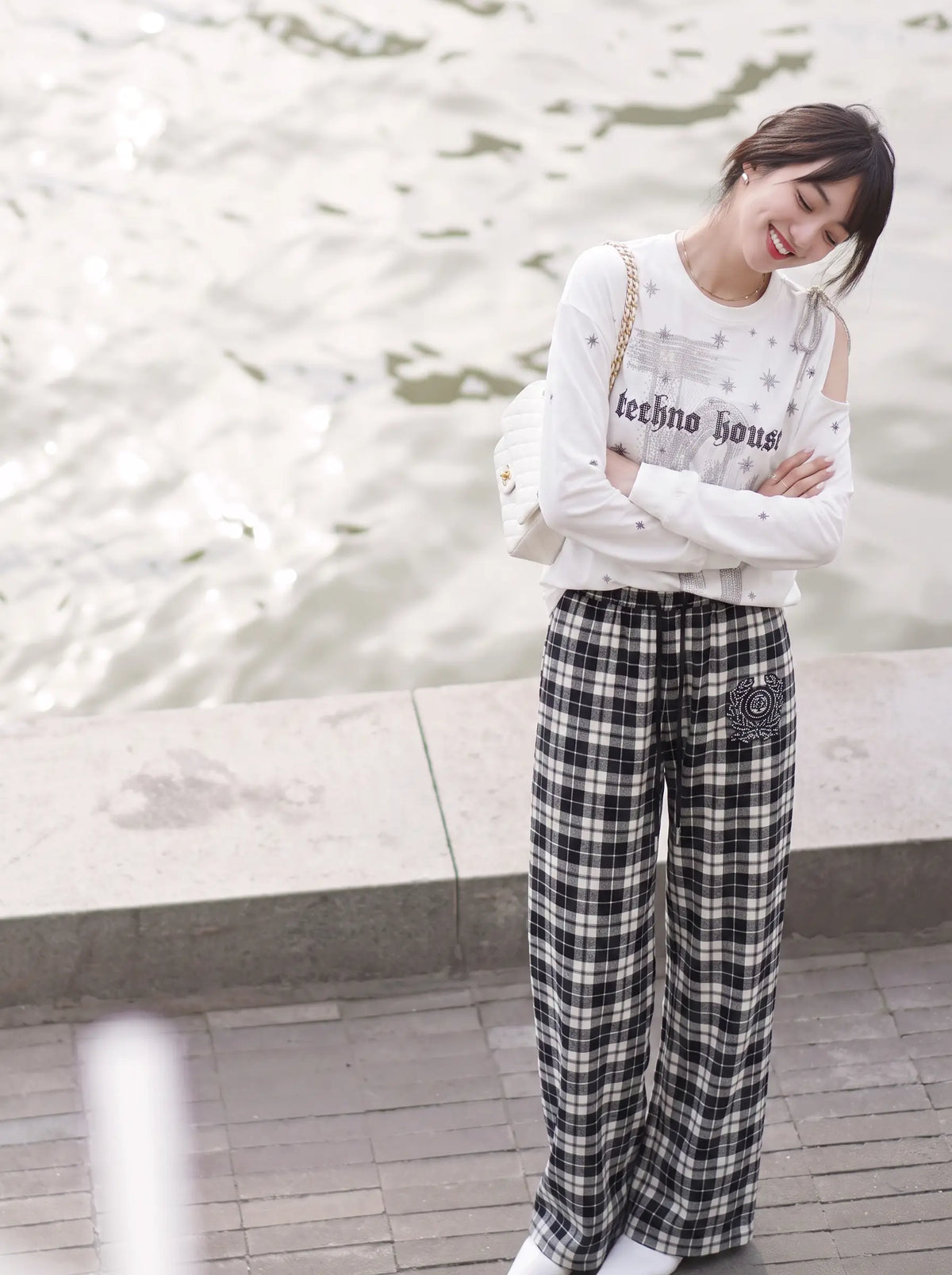 A woman stands outdoors near water, wearing a white long-sleeve top with 'leethno house' printed on the front and black-and-white plaid wide-leg pants. TCH Starry Western Graphic White Long Sleeve Top