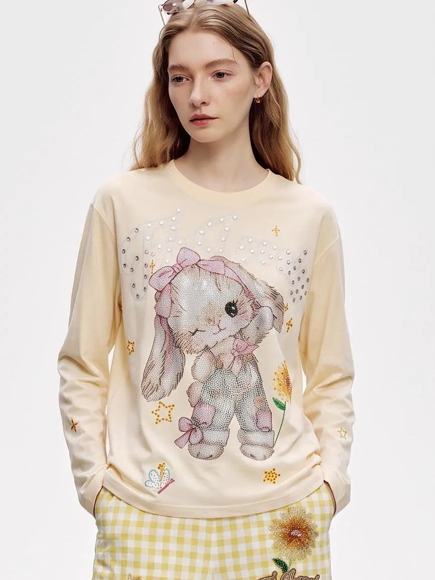 A young woman wearing a cream-colored long-sleeve sweatshirt with a large pink and gray bunny graphic, decorated with rhinestones and gold star accents, paired with yellow and white gingham pants. TCH Sweet Bunny Rhinestone Graphic Long Sleeve Top