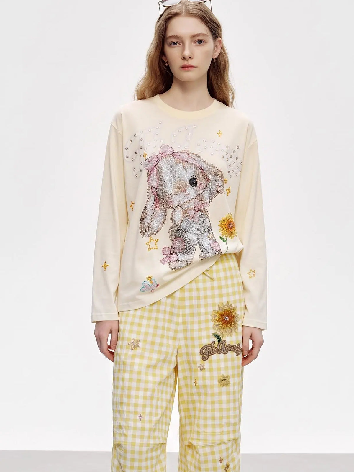 A model wearing a cream-colored long-sleeve t-shirt with a bunny graphic and yellow-and-white gingham pants with a sunflower patch. TCH Sweet Bunny Rhinestone Graphic Long Sleeve Top