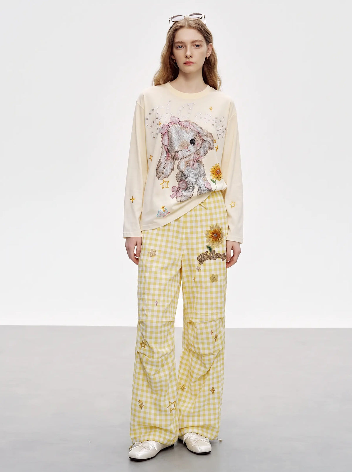 A model stands against a light gray studio background wearing a cream long-sleeve top with a bunny graphic and yellow-and-white gingham pants with small embroidered details. TCH Sweet Bunny Rhinestone Graphic Long Sleeve Top