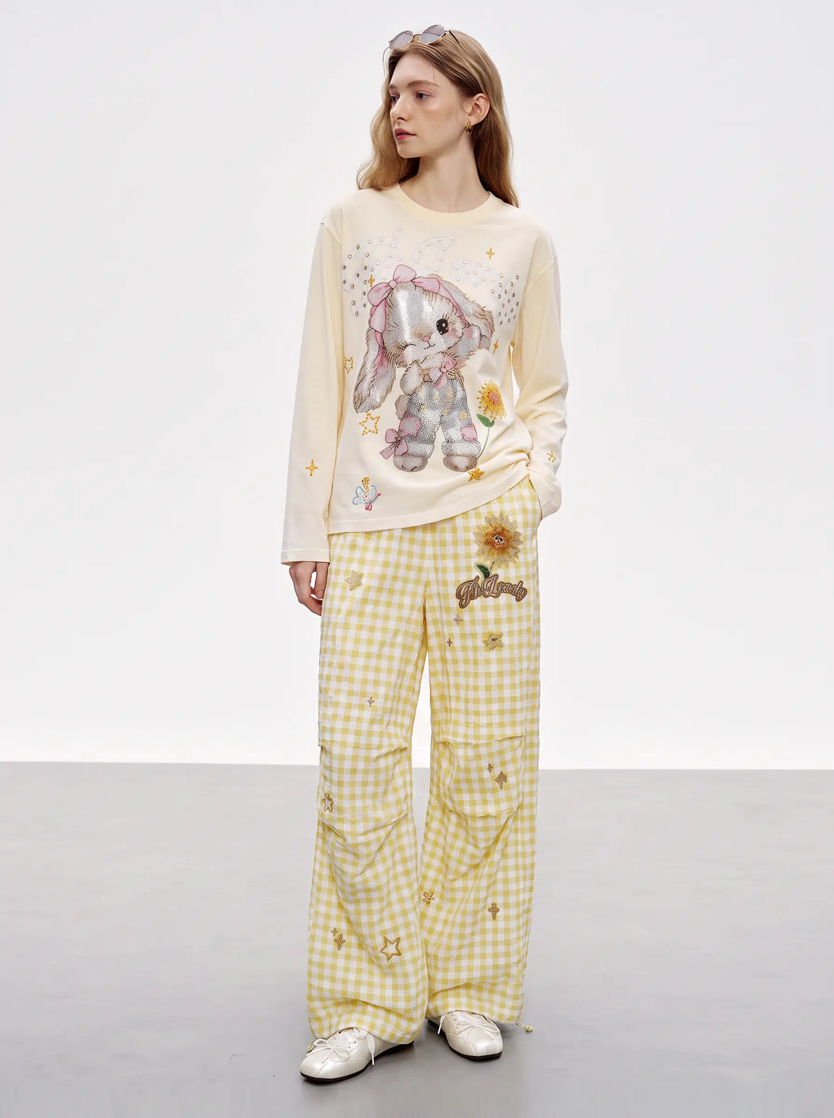 A model stands in a studio wearing a cream long-sleeve top with a bunny graphic and yellow gingham pants with sunflower prints. TCH Sweet Bunny Rhinestone Graphic Long Sleeve Top