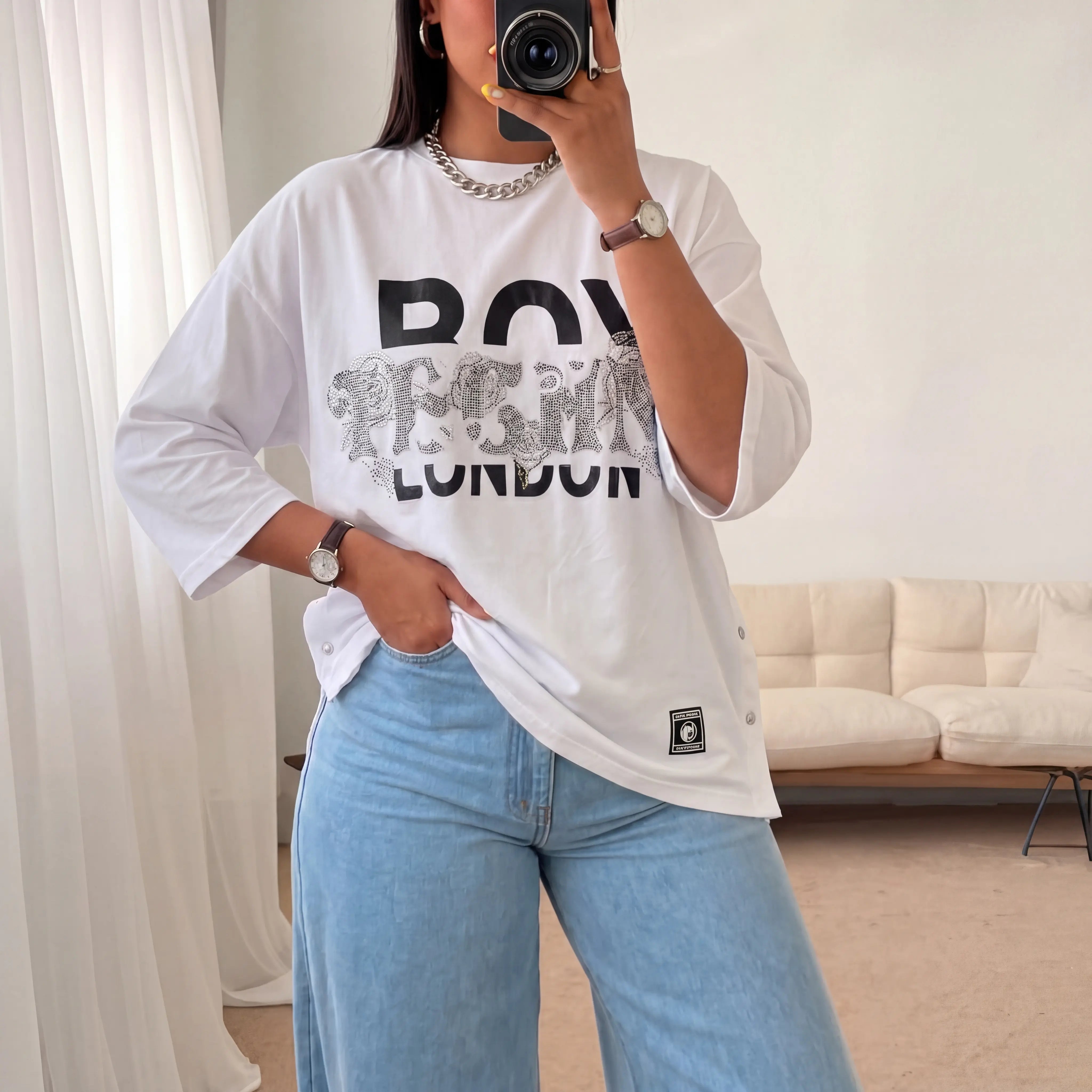 Person wearing a white oversized t-shirt with text and blue jeans in a room. TCH TECHNO Rhinestone Layered Typography T-Shirt