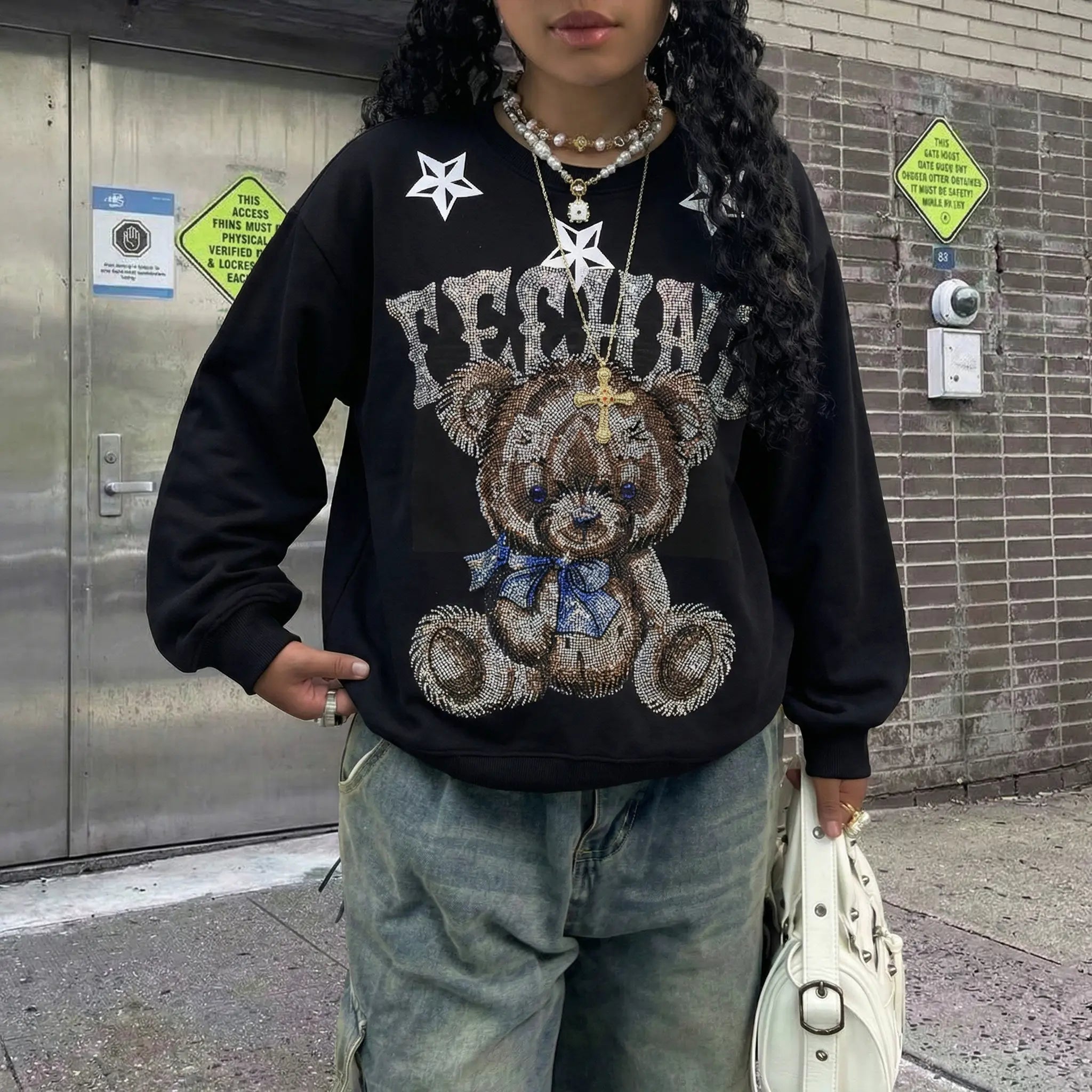 Person wearing a black sweatshirt with a teddy bear graphic and text, standing against a brick wall. TCH TECHNO Rhinestone Teddy Sweatshirt