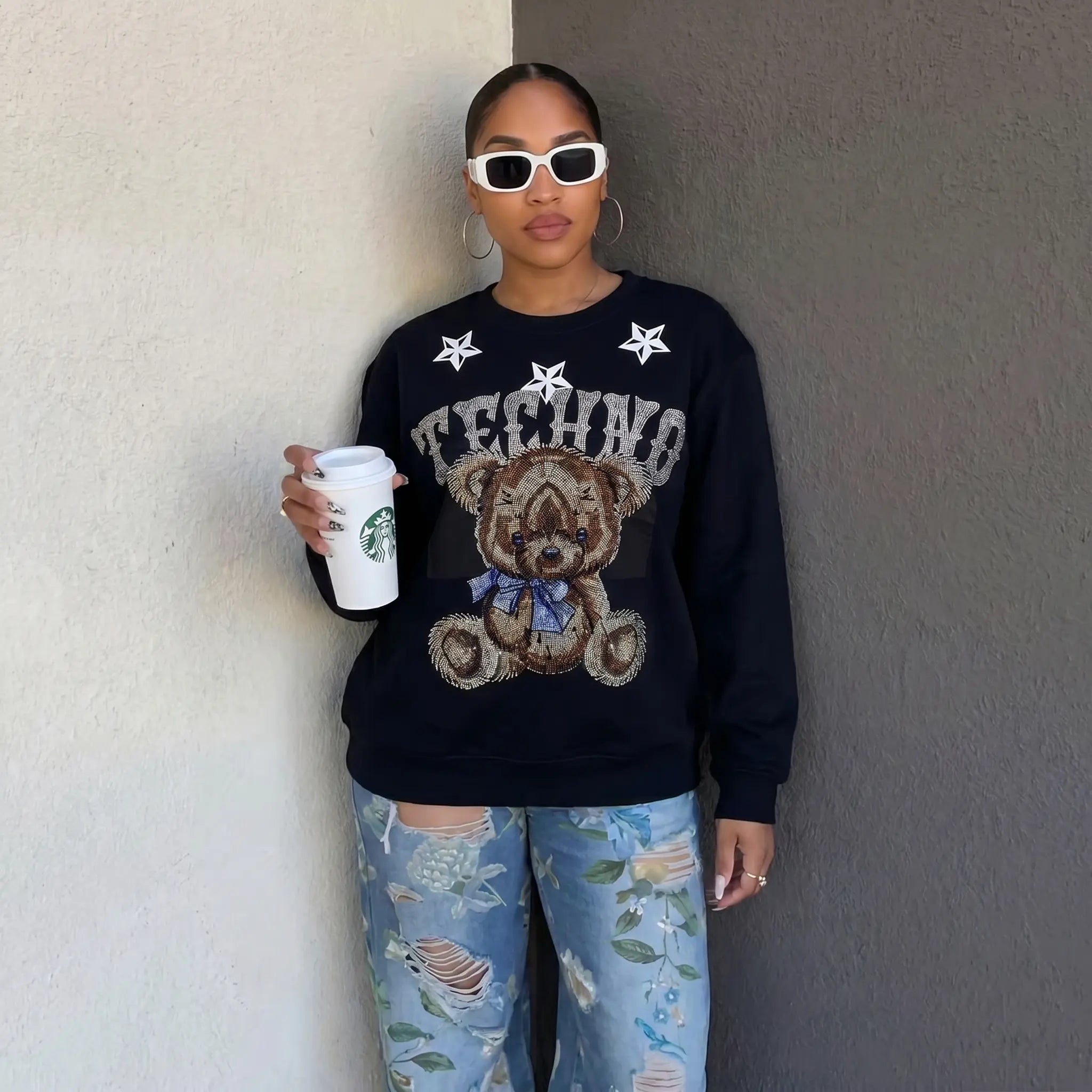 Person wearing a black sweater with a teddy bear graphic and holding a Starbucks cup against a plain wall. TCH TECHNO Rhinestone Teddy Sweatshirt