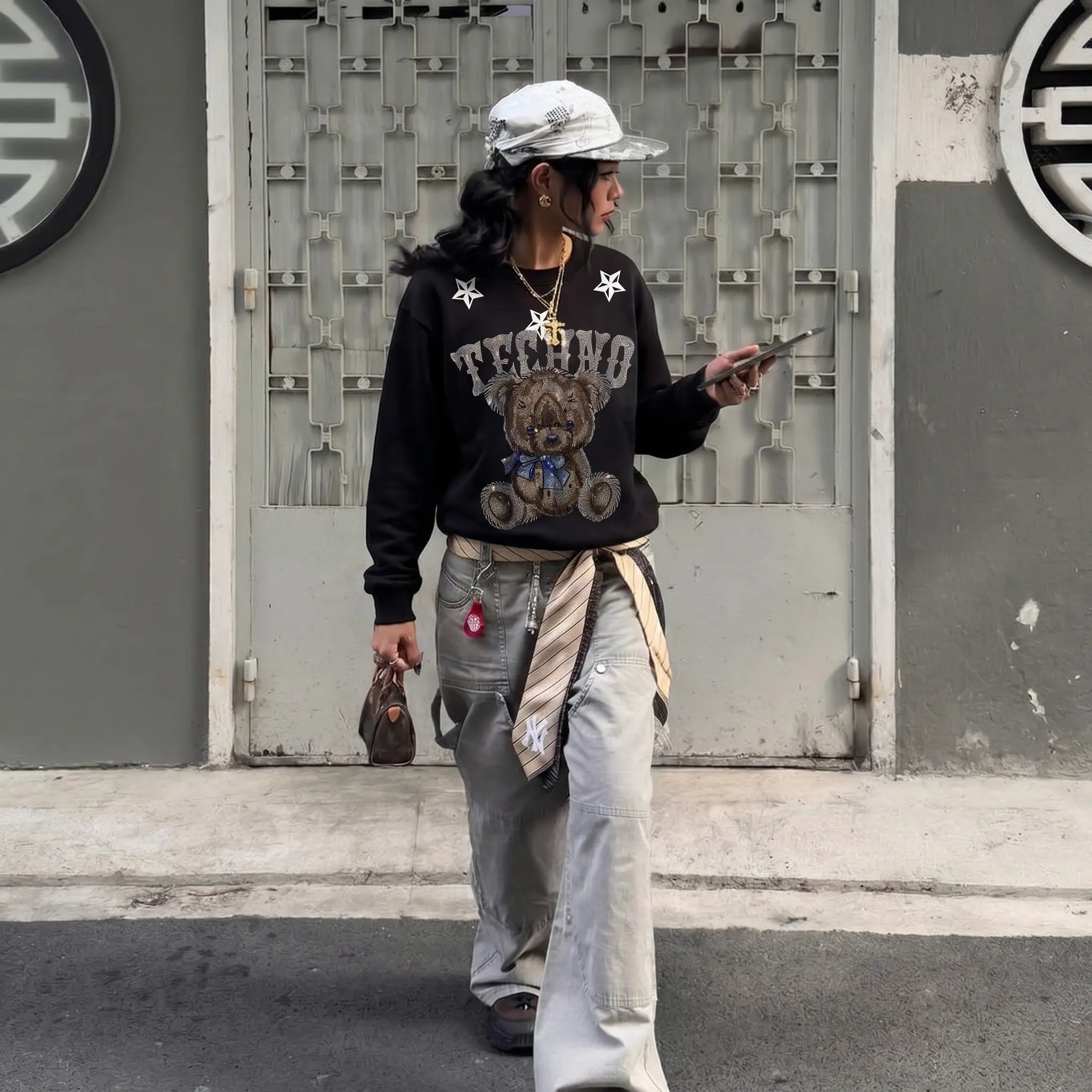 Person wearing a black sweater with a graphic design, light-colored pants, and a white cap, standing on a street. TCH TECHNO Rhinestone Teddy Sweatshirt