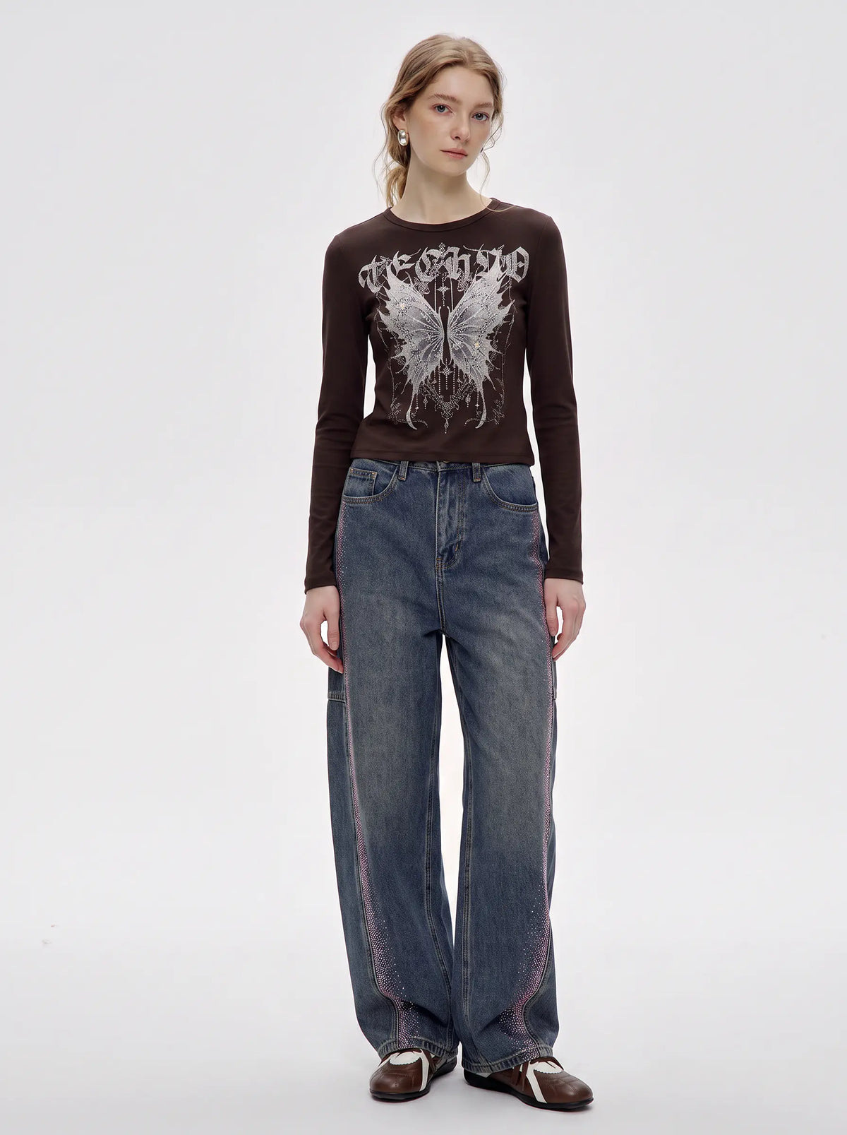 A model stands against a light gray studio background wearing a dark brown long-sleeve graphic t-shirt with a white distressed butterfly design and the word 'DEGENERATE' across the chest, paired with wide-leg blue jeans and brown leather shoes. TCH Techno Butterfly Starburst Graphic Long Sleeve Top