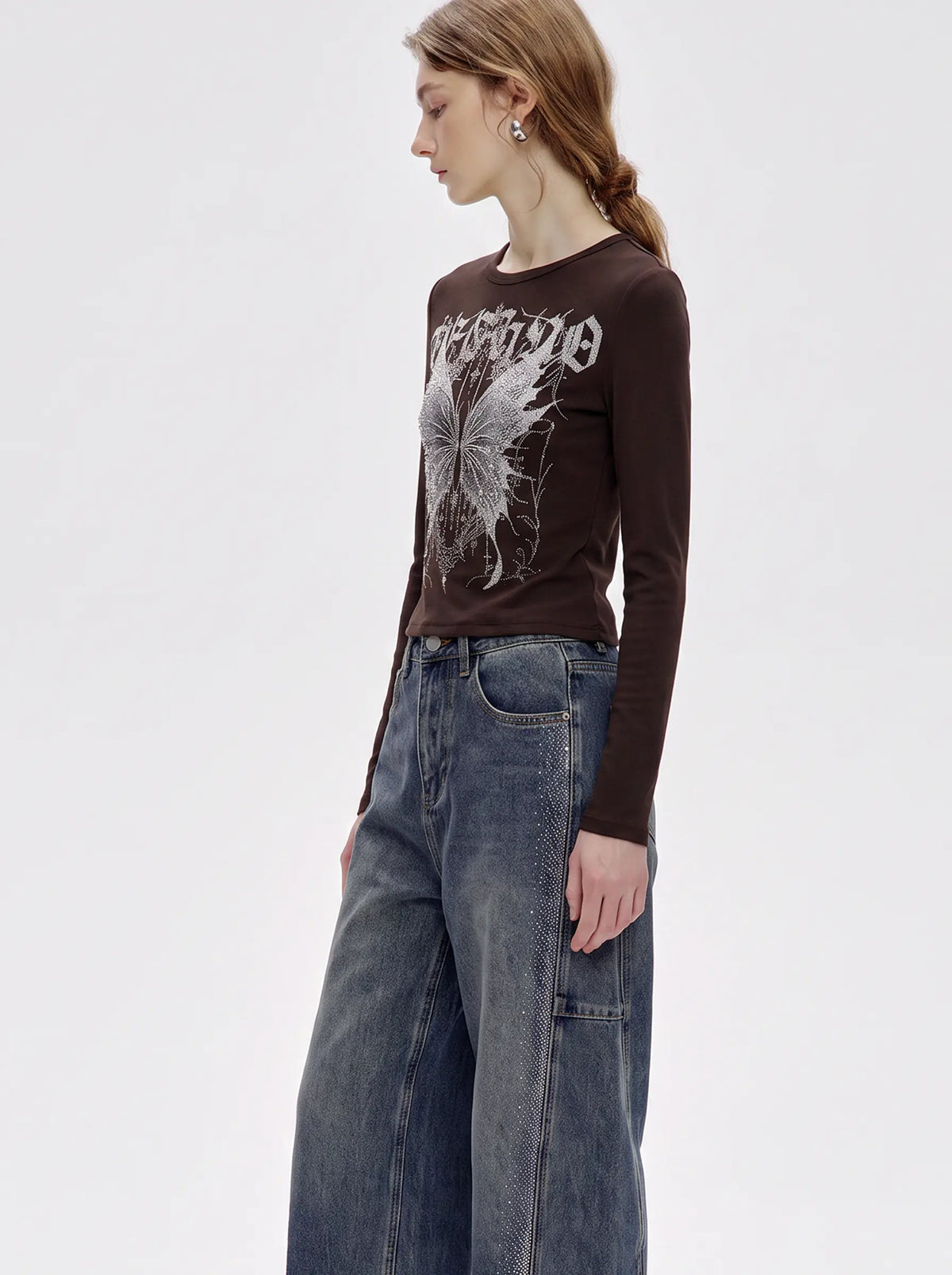 A woman modeling a dark brown long-sleeve t-shirt with a white graphic print on the front, paired with wide-leg blue jeans, against a plain light gray background. TCH Techno Butterfly Starburst Graphic Long Sleeve Top