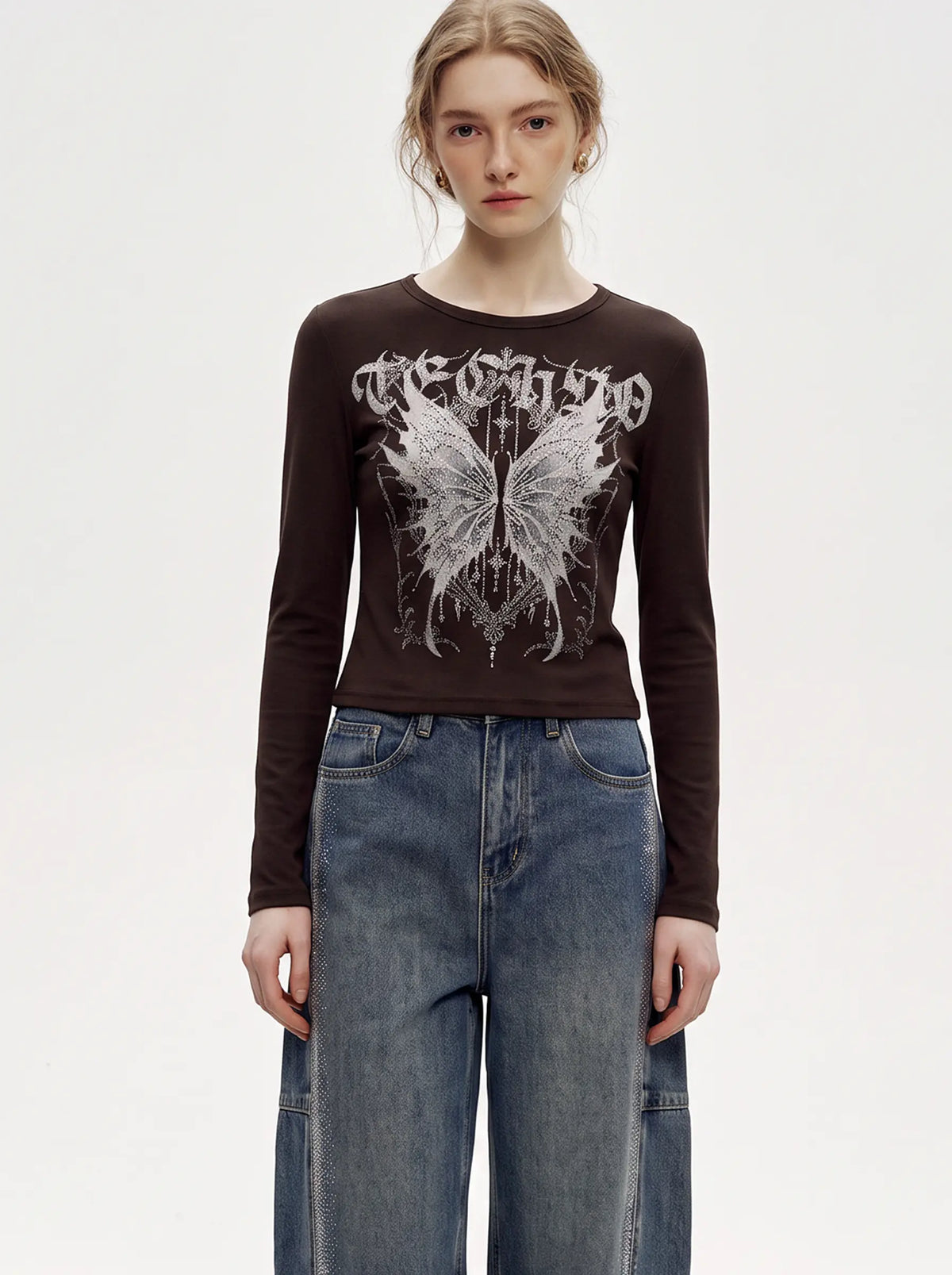 A model wearing a dark brown long-sleeve crop top with a white gothic-style graphic of a butterfly and the word 'Techno' on the front, paired with high-waisted blue jeans, against a plain white studio background. TCH Techno Butterfly Starburst Graphic Long Sleeve Top