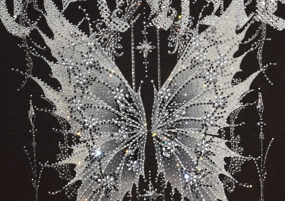 A close-up of a black garment covered with intricate silver rhinestone and crystal embellishments, forming a symmetrical wing-like pattern with dangling sparkly details. TCH Techno Butterfly Starburst Graphic Long Sleeve Top