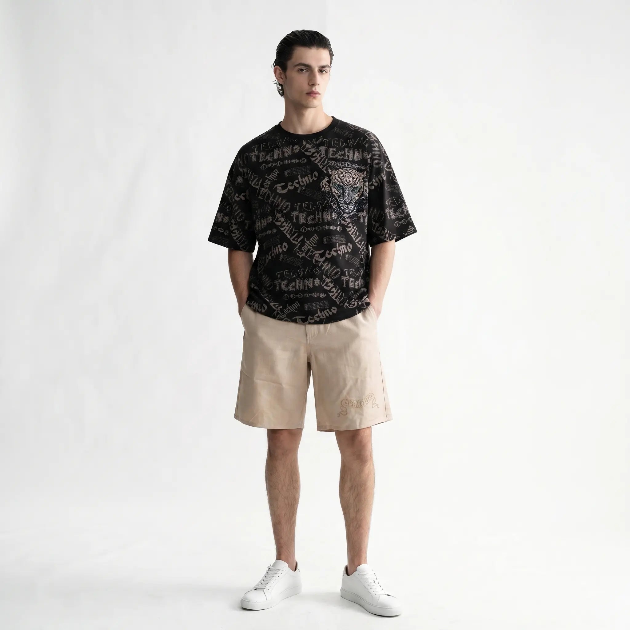 A man modeling a black graphic t-shirt with a leopard print and 'TECHNO' text, paired with beige shorts and white sneakers, standing against a white studio background. TCH Techno Graffiti Rhinestone Skull Oversized T-Shirt