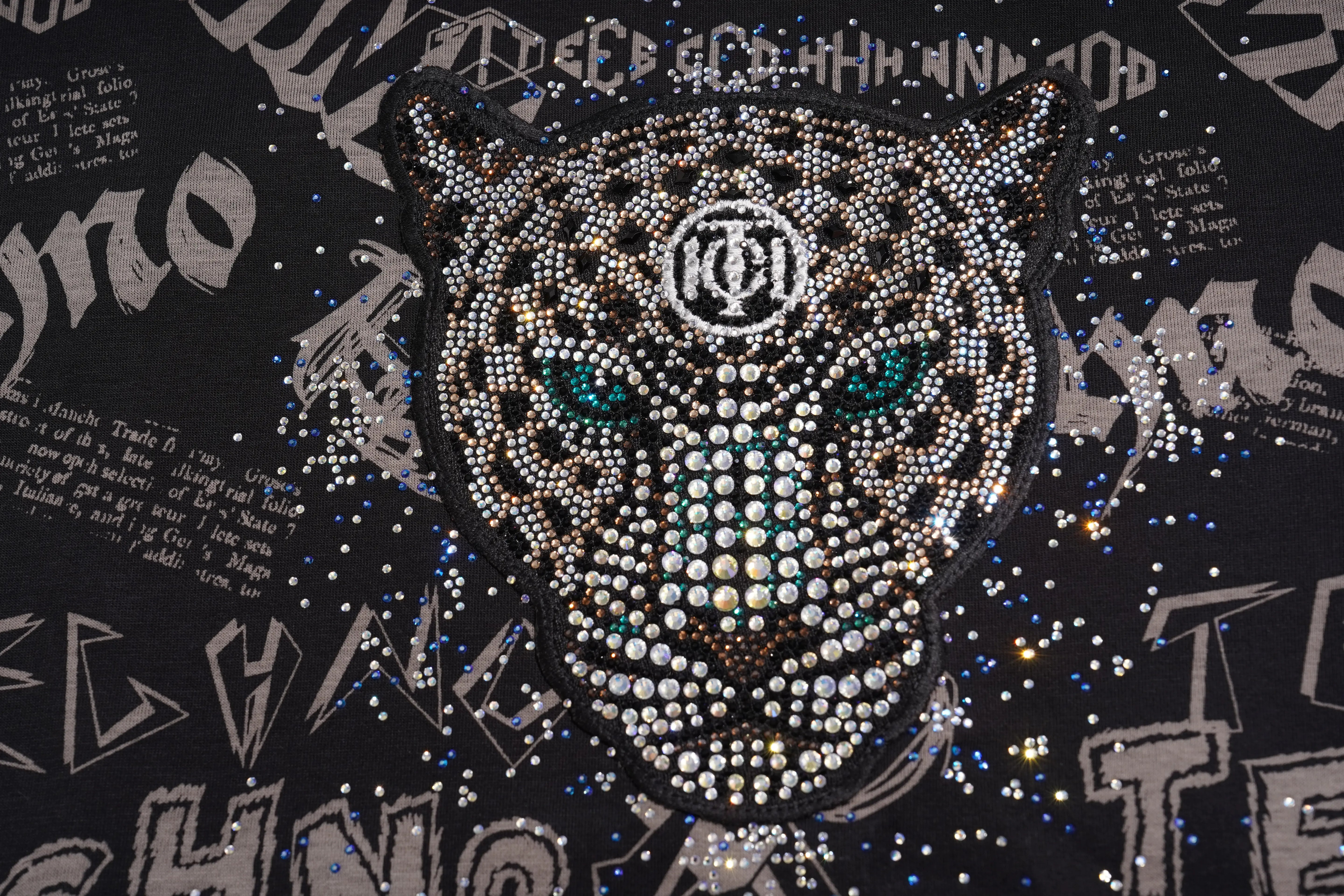 A close-up of a black garment featuring a large rhinestone-embellished panther head with teal eyes and a circular logo on the forehead, surrounded by scattered rhinestones and graffiti-style lettering. TCH Techno Graffiti Rhinestone Skull Oversized T-Shirt