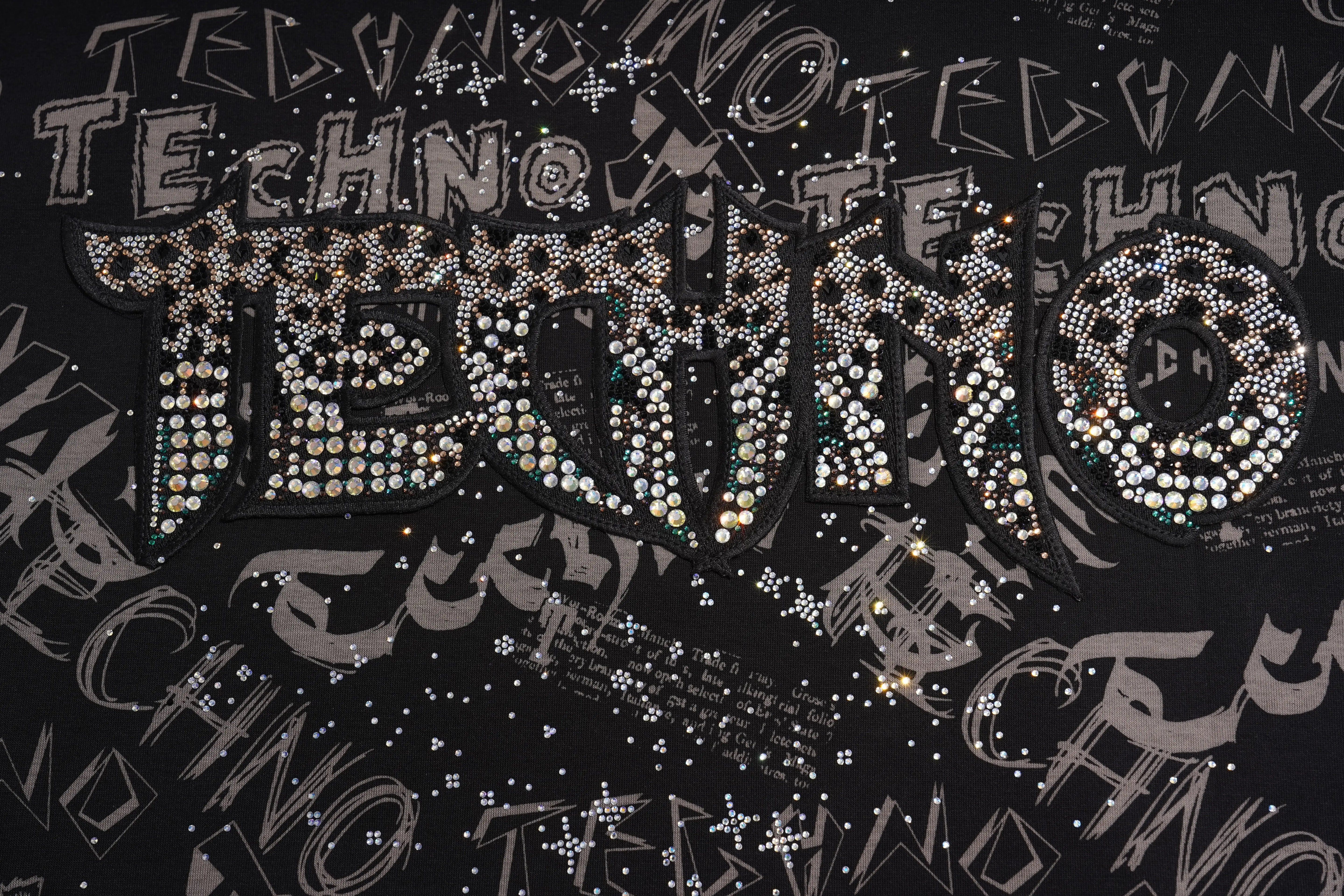 A close-up of a black garment covered with layered, rhinestone-embellished lettering spelling TECHNO, with additional scattered rhinestone text and small reflective embellishments across the surface. TCH Techno Graffiti Rhinestone Skull Oversized T-Shirt