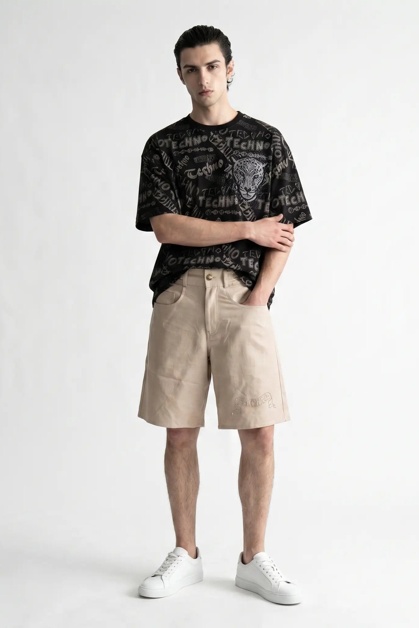A male model stands against a light gray studio background wearing a black graphic t-shirt, beige shorts, and white sneakers. TCH Techno Graffiti Rhinestone Skull Oversized T-Shirt