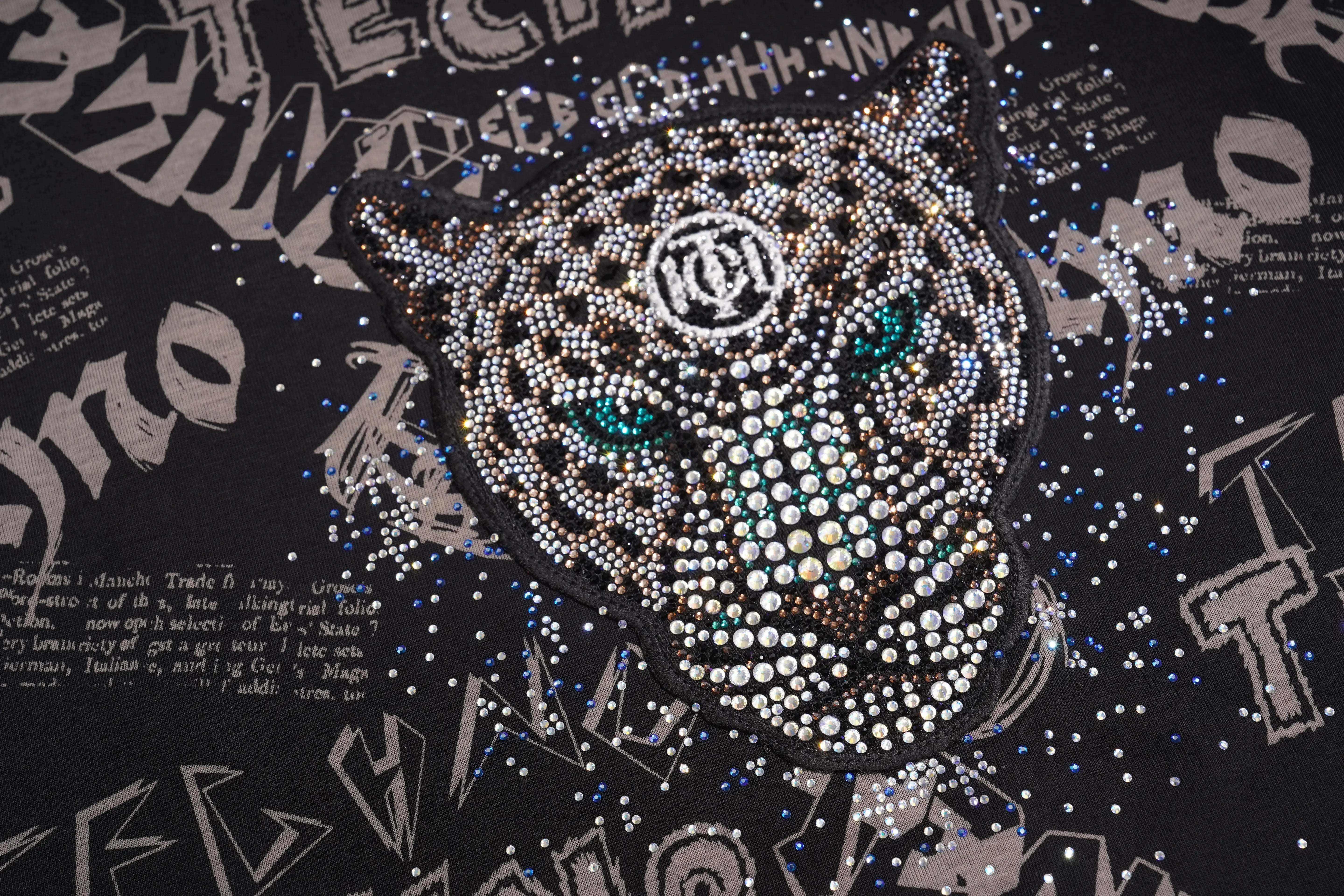 A close-up of a black garment featuring a large panther head design made of rhinestones, with turquoise-colored eyes and a circular monogram logo on the forehead, surrounded by abstract graphic patterns. TCH Techno Graffiti Rhinestone Skull Oversized T-Shirt