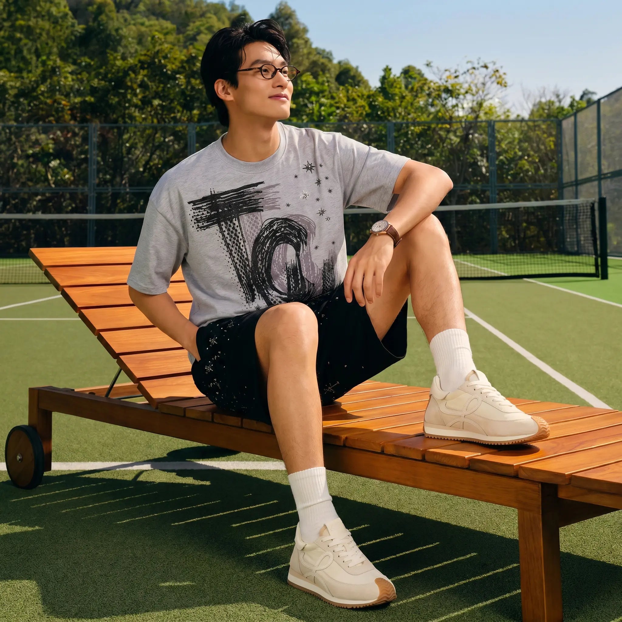 A man wearing glasses sits on a wooden lounge chair on a tennis court, wearing a light gray graphic t-shirt, black shorts, white socks, and white sneakers.TCH Techno House Abstract Graphic Oversized T-Shirt