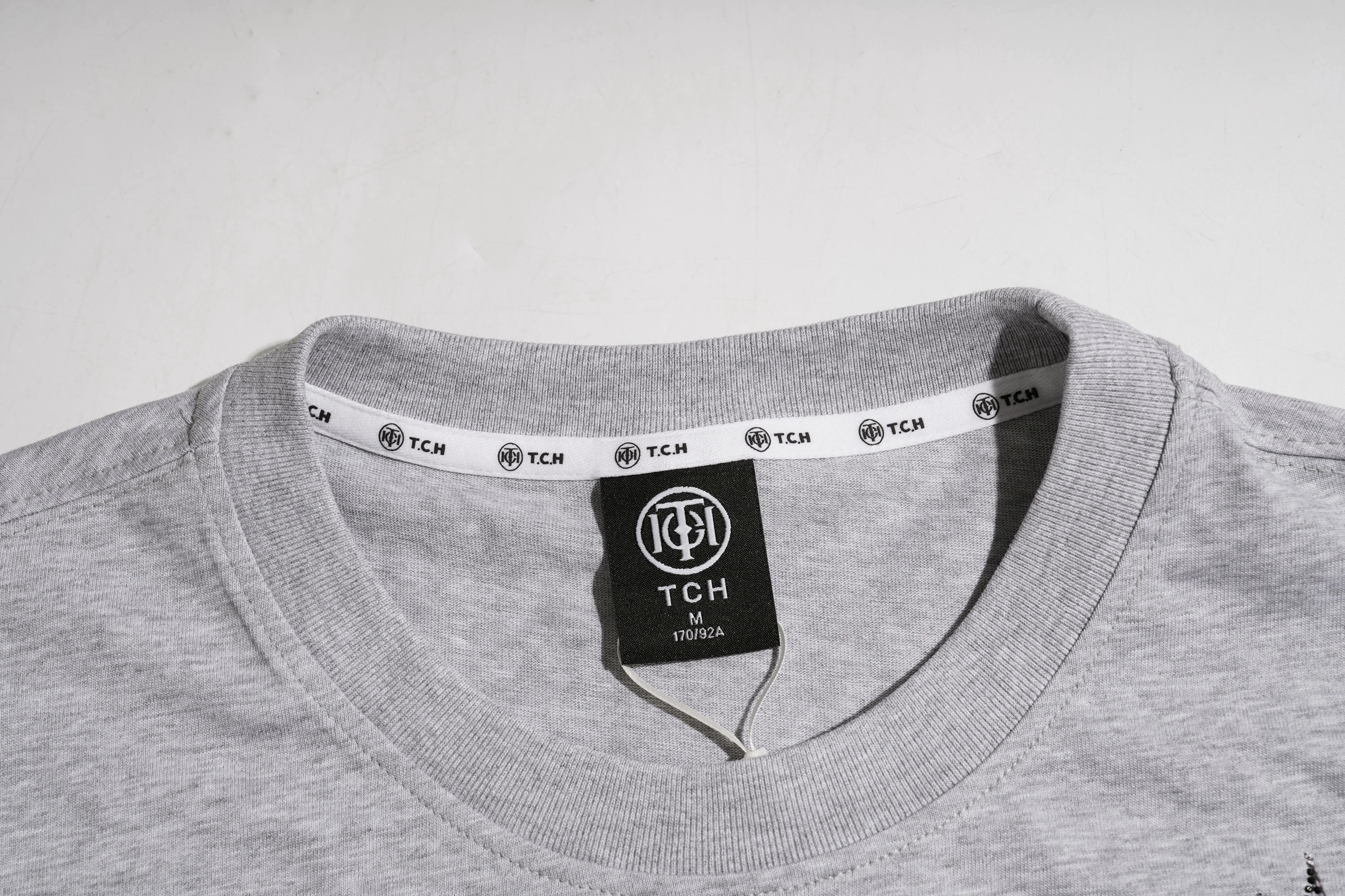 Close-up of a light gray heather crewneck t-shirt showing the inner collar with a white printed TCH brand band and a black TCH hangtag. TCH Techno House Abstract Graphic Oversized T-Shirt