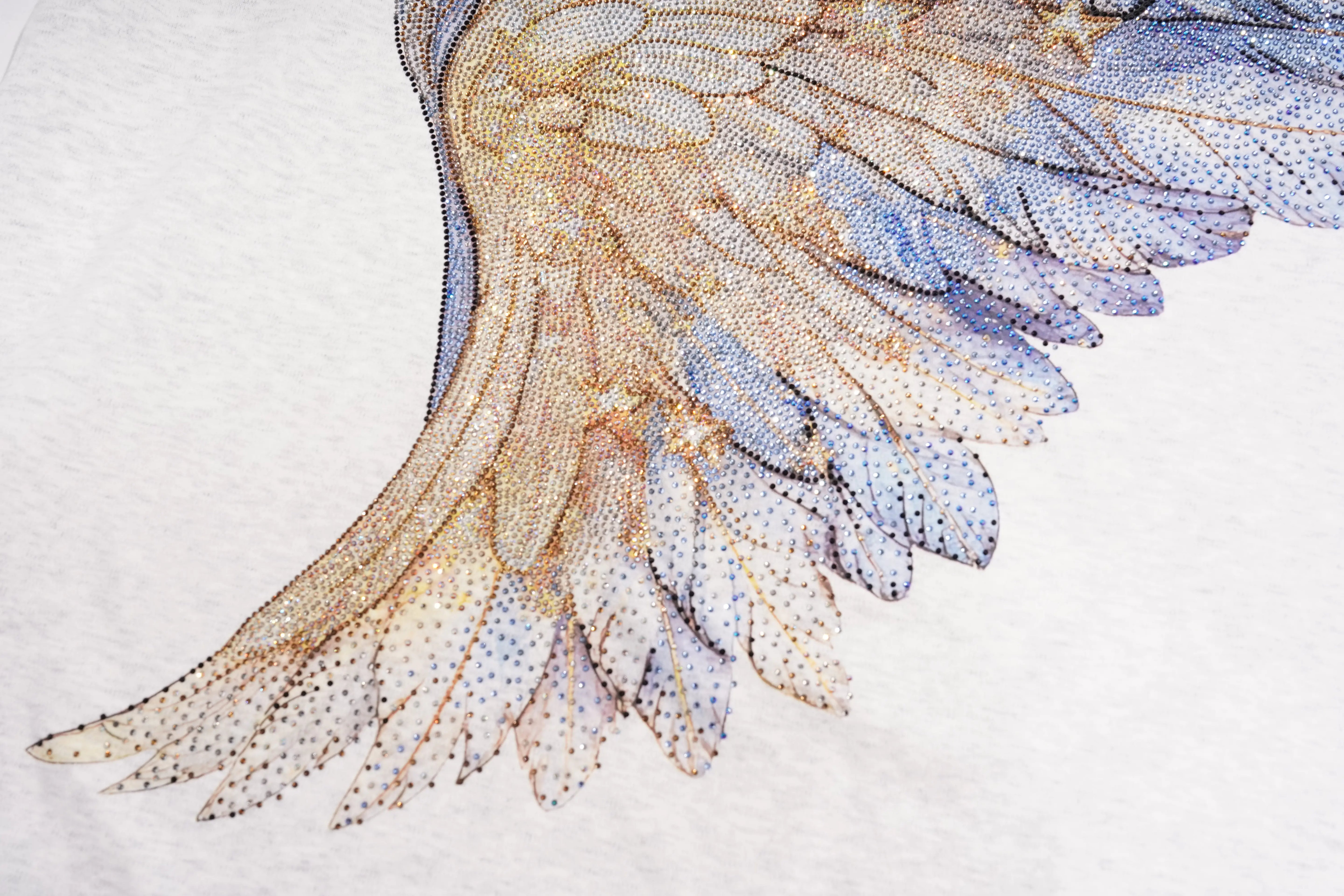 A close-up of a detailed wing design made from many small rhinestones, showing layered feathers in warm gold, beige, and cool blue tones on a white background. TCH Techno House Wing Graphic Oversized T-Shirt