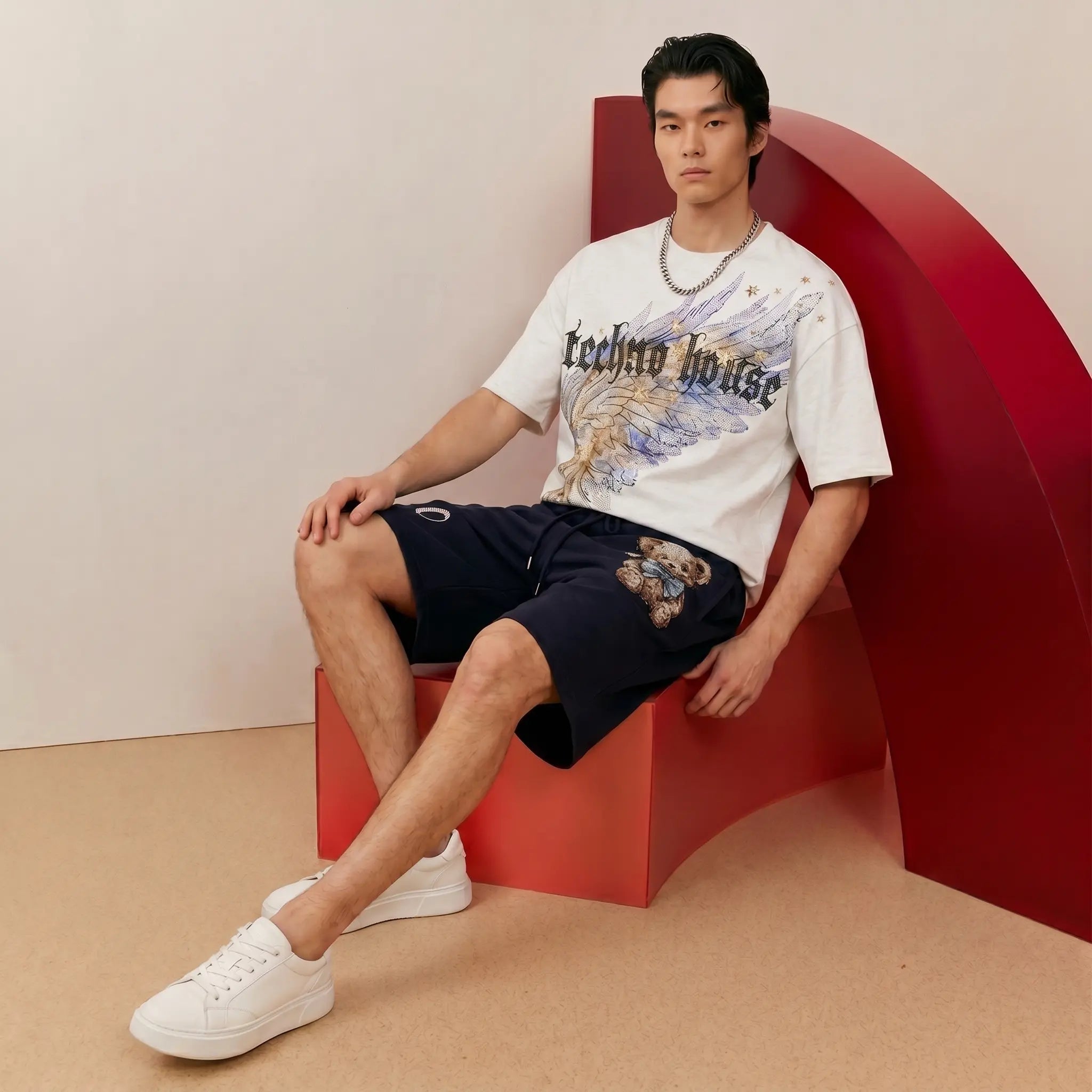 A man models a white graphic t-shirt with 'Techno house' text and a blue/gold eagle design, paired with navy shorts featuring a tiger graphic and white sneakers, seated on a red geometric bench. TCH Techno House Wing Graphic Oversized T-Shirt