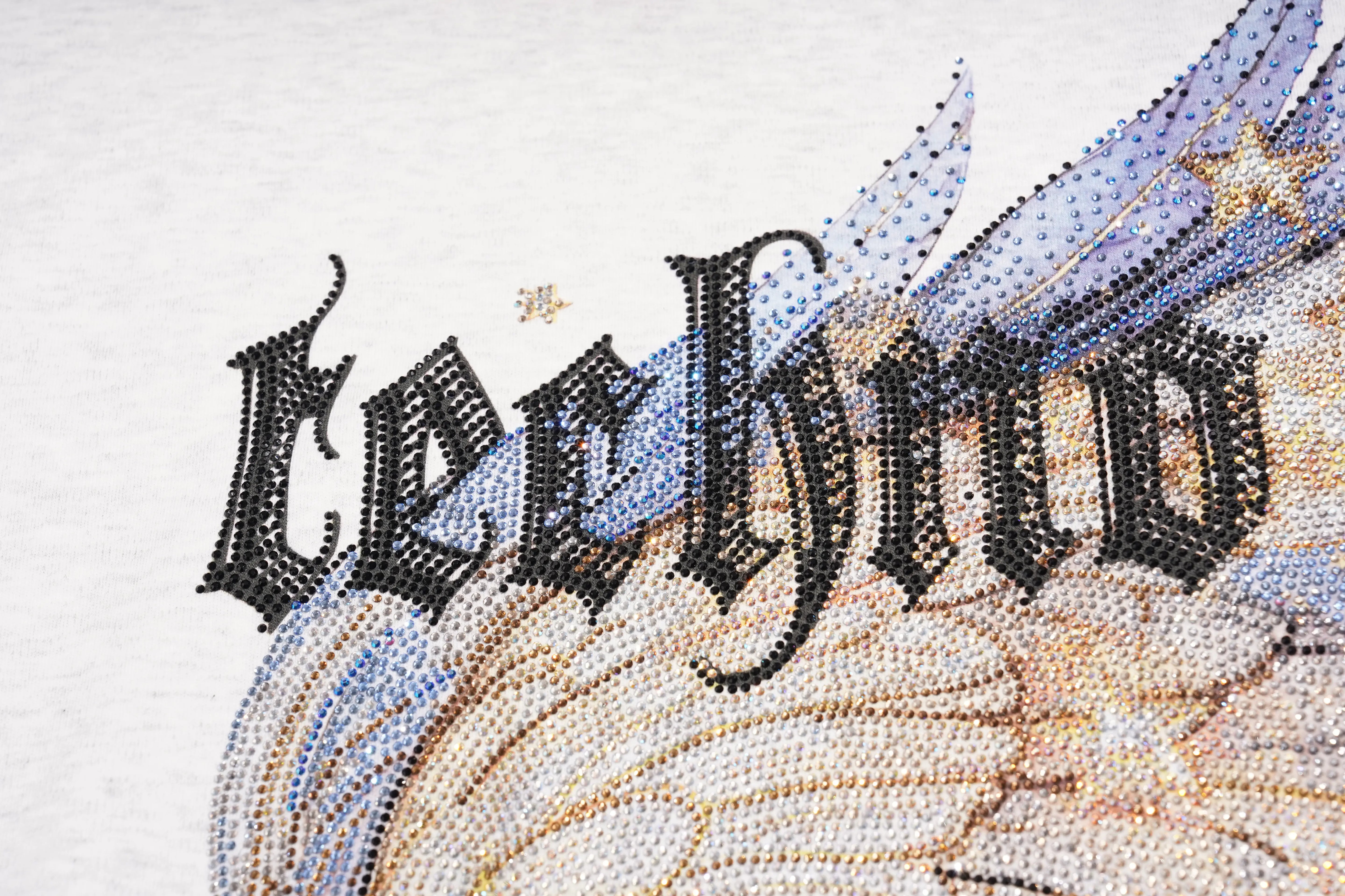 A close-up of a textured fabric featuring the word 'Lechino' in a black gothic font, overlaid with blue and gold glittery wing-like patterns on a white background. TCH Techno House Wing Graphic Oversized T-Shirt